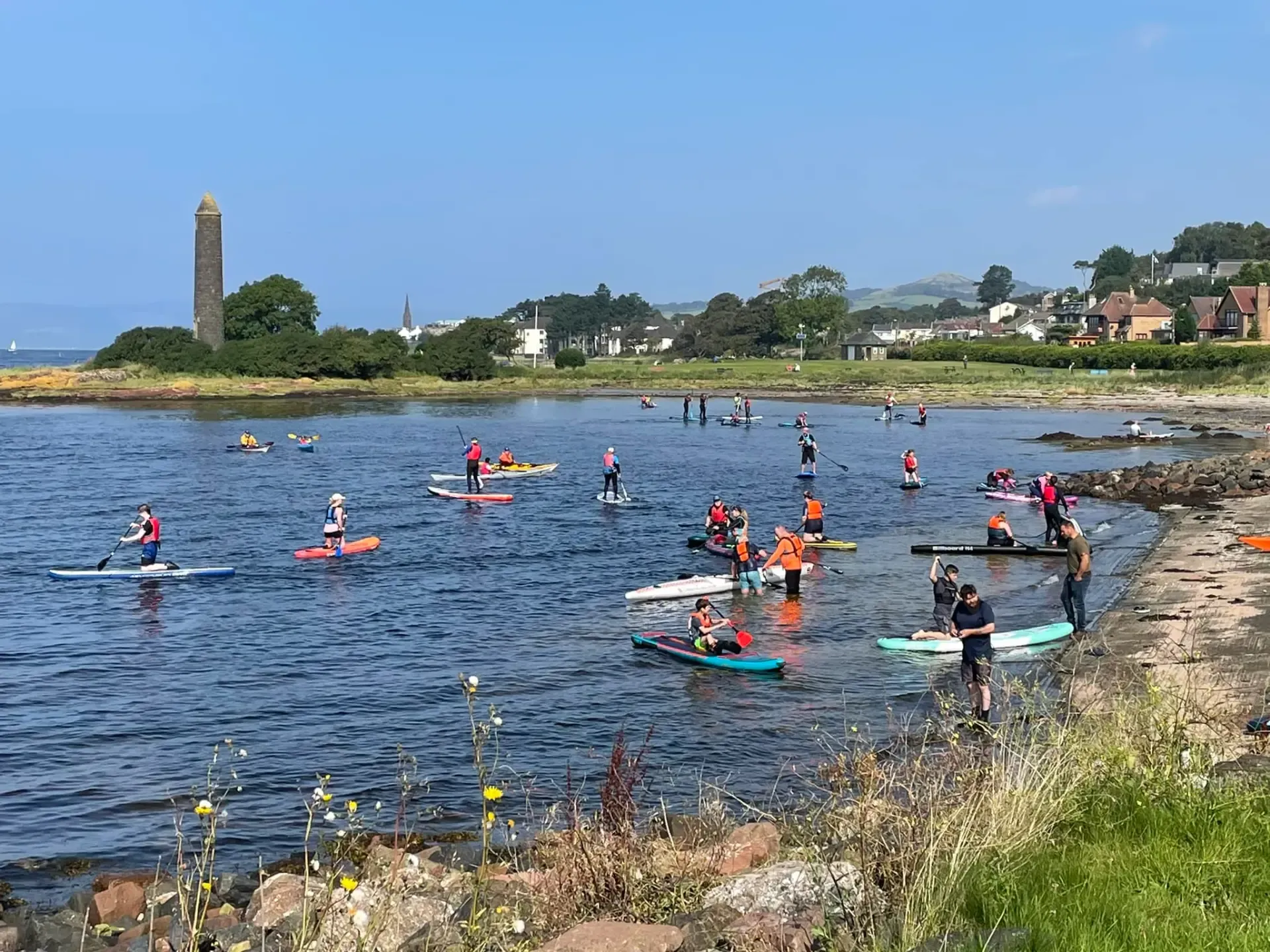People paddleboard in a bay with a stone tower on a sunny day. Some wear orange vests; water is calm.