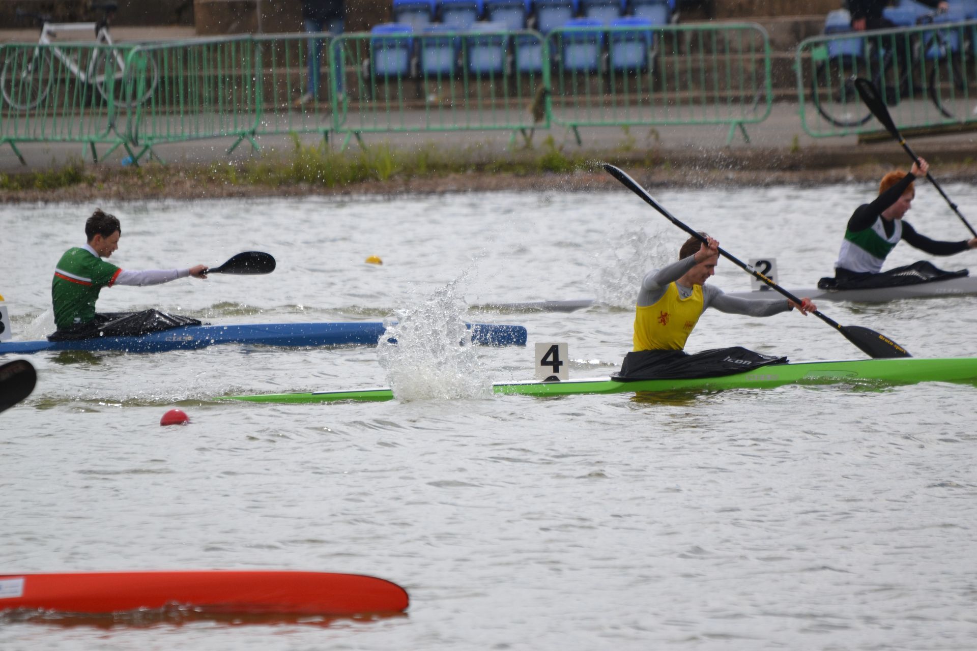 Canoe racers in a lake. A man in a yellow shirt paddles, creating water spray. Others compete nearby in green and white uniforms.