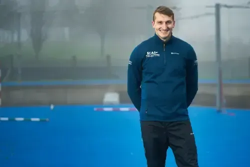 Man in a blue jacket smiles, standing with hands behind his back on a blue sports field; a blurred background hints at an outdoor setting.