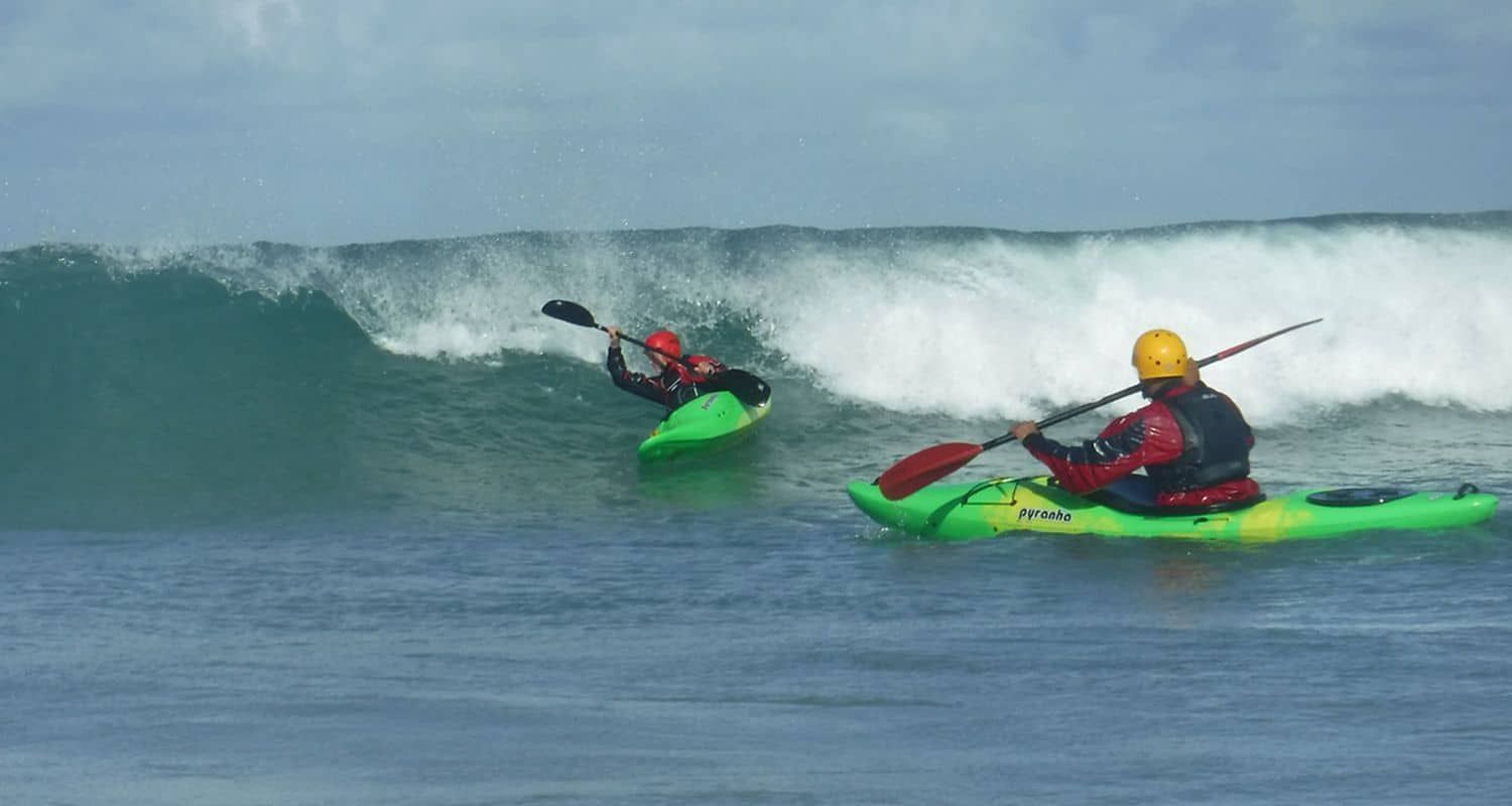 Two kayakers riding waves in the ocean. One flips upside down, while the other paddles forward. Bright blue sea and sky.