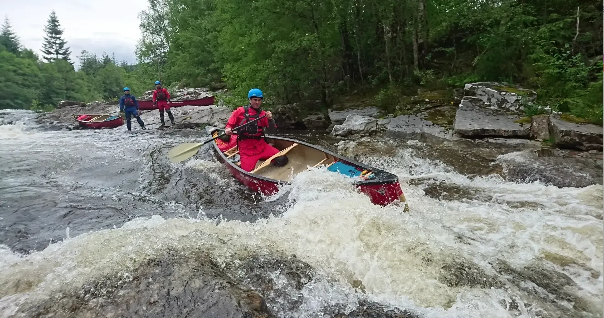 A person in a red suit paddles a canoe through whitewater rapids. Other people and canoes are visible on the riverbank.
