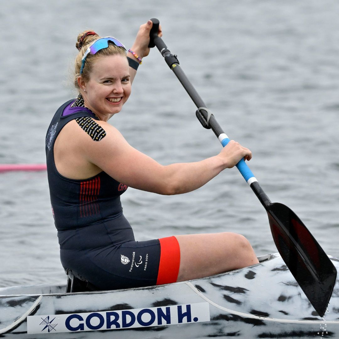 A female athlete in a kayak paddles on a lake, smiling. She wears athletic clothing with kinesiology tape.