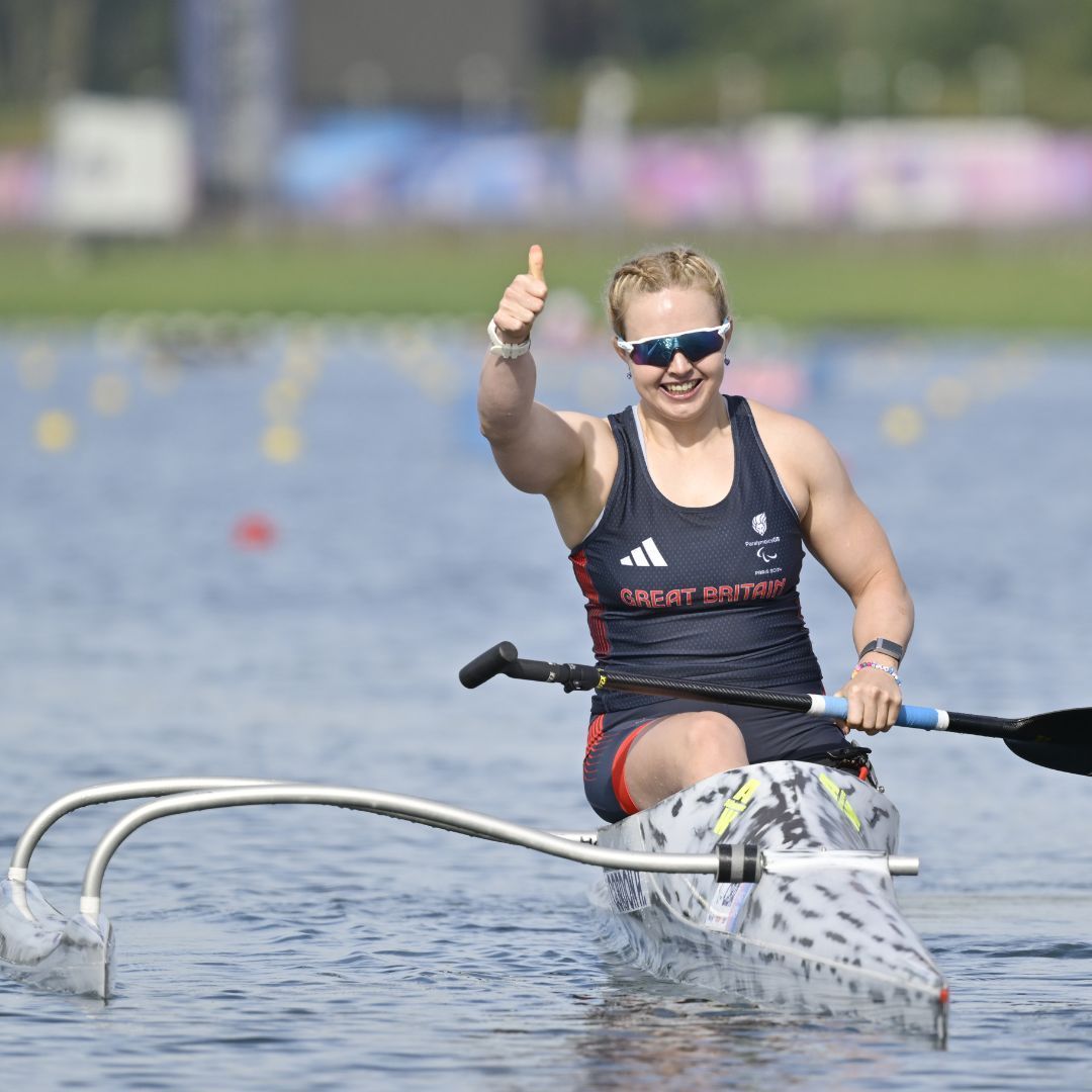 Female para-canoeist in a Great Britain uniform giving a thumbs up, smiling, on a lake.