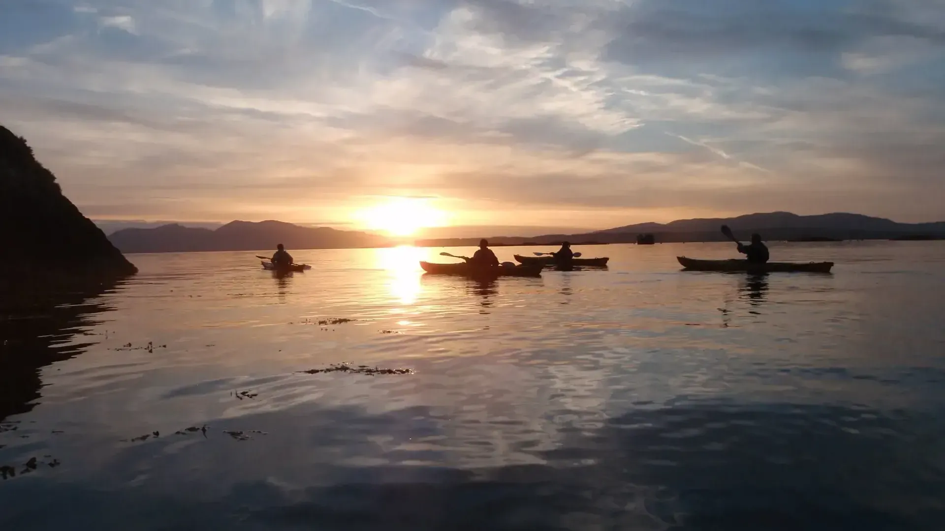 Kayakers on calm water at sunset, silhouetted against orange sky.