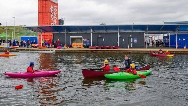 Kayakers and canoeists paddling on a lake near a building with a large orange structure in the background. People are present on the dock.
