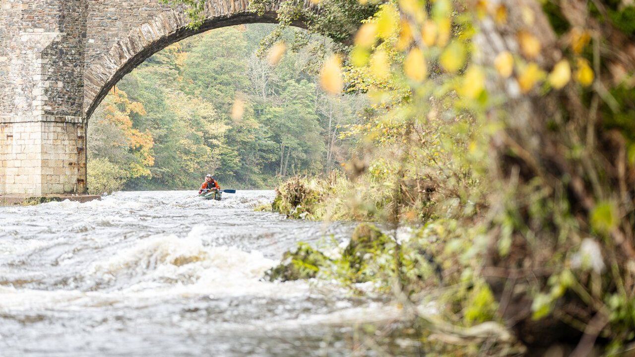 A person kayaks under an arched stone bridge on a rushing river surrounded by autumn foliage.