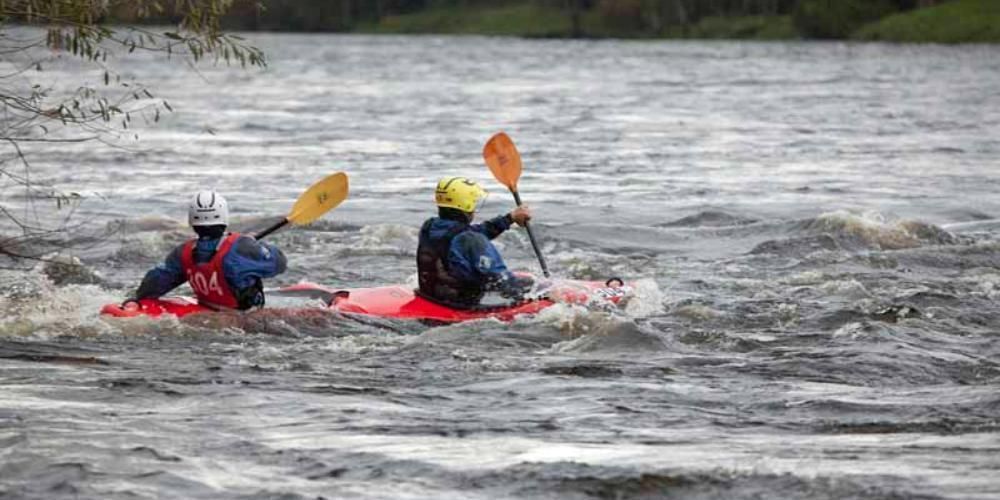 Two kayakers navigating rough, grey water in red kayaks. The kayakers wear helmets and use paddles.