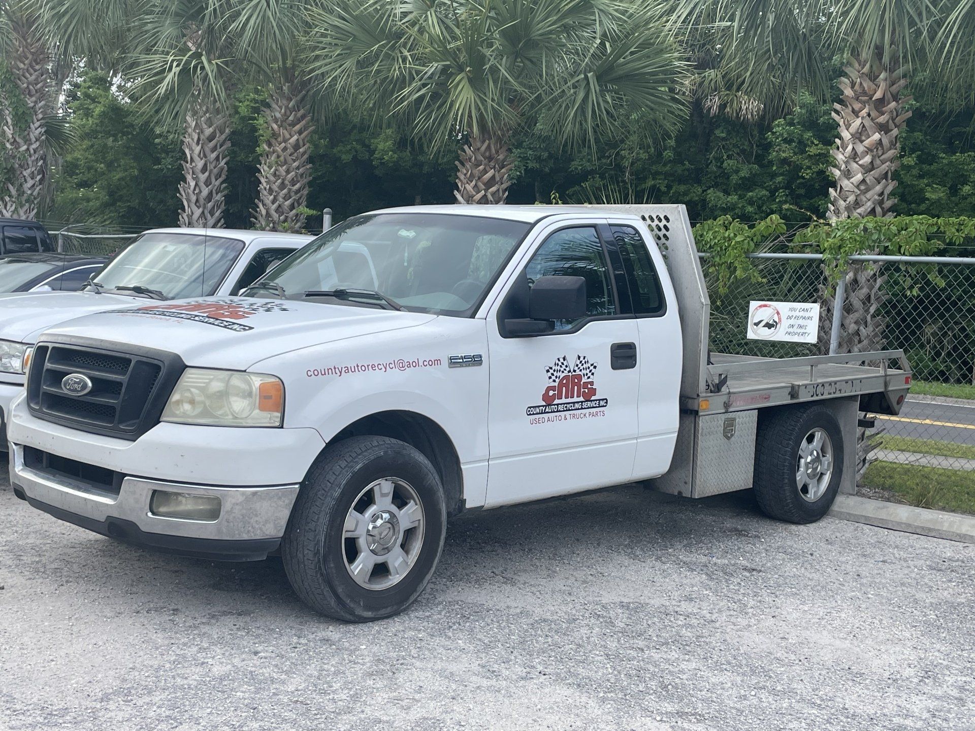 White Ford F-150 flatbed truck parked on gravel with logo on door. Green palm trees in background.