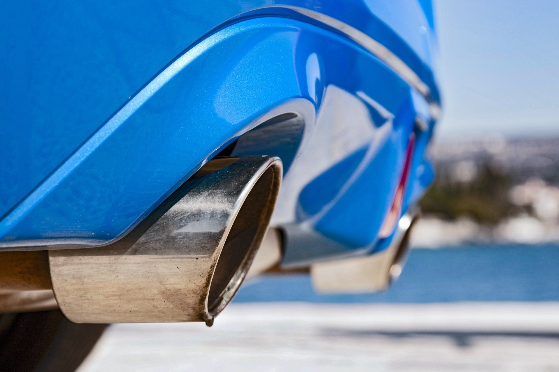 Close-up of blue car's shiny exhaust pipes, set against a blurred background of water and sky.