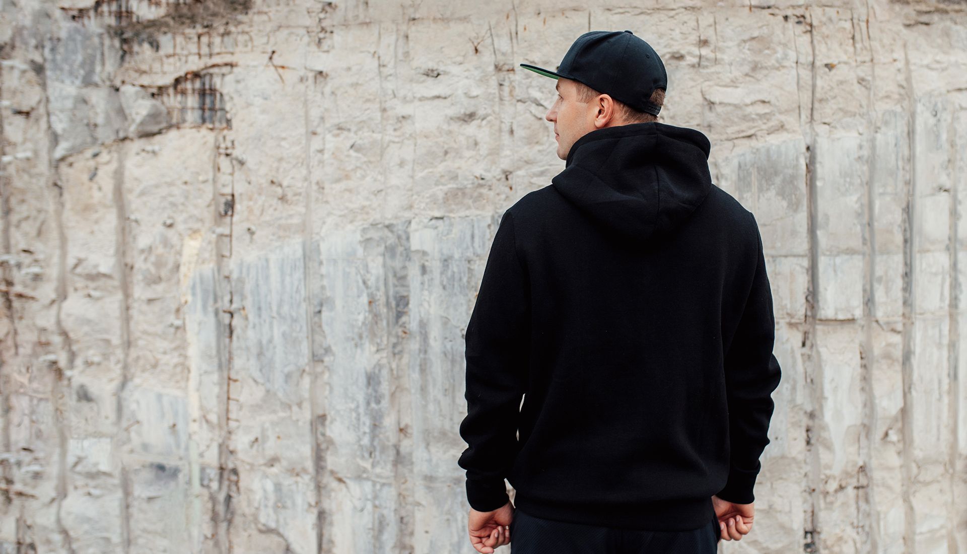 Man in black hoodie and cap, standing in front of a textured concrete wall, looking away.
