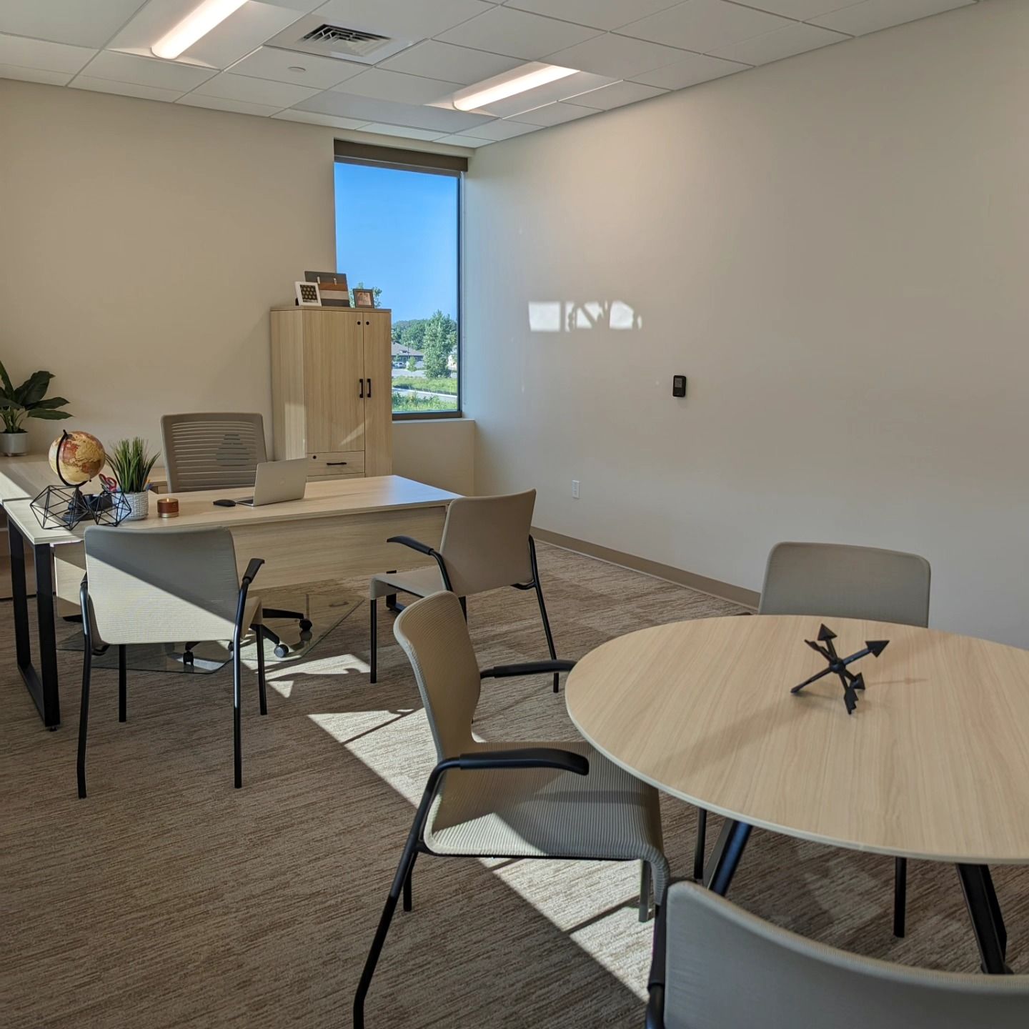 Office space with a large desk, chairs, and a round table. Sunlight streams in from a window.