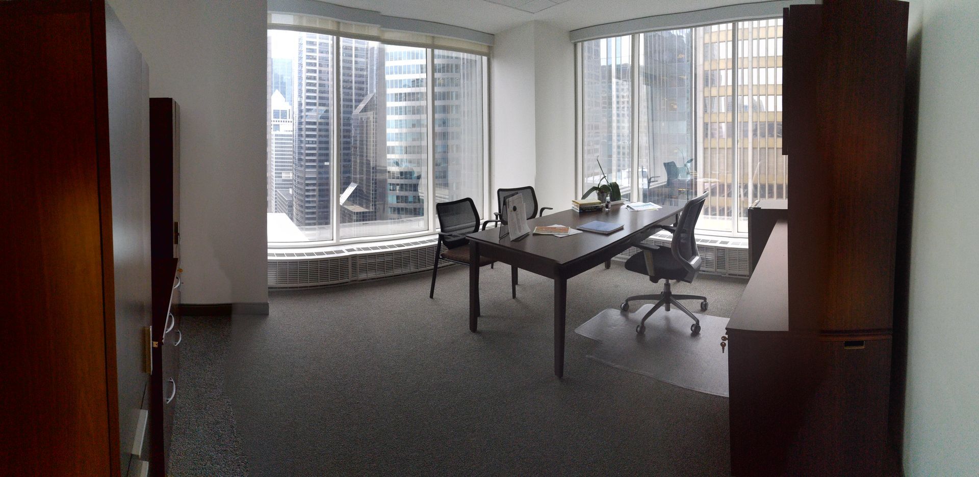 Empty office with large windows overlooking city buildings; a long table and chairs are in the center.