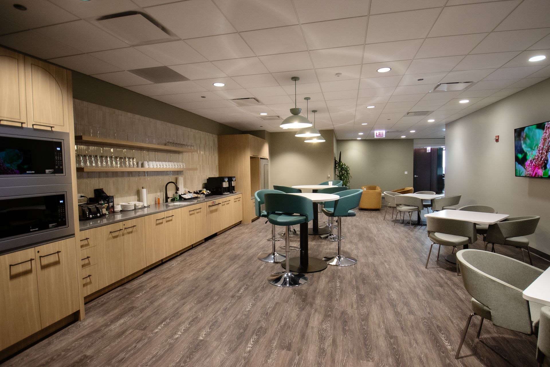 Office breakroom with kitchen, seating areas, light wood cabinets, and gray flooring.