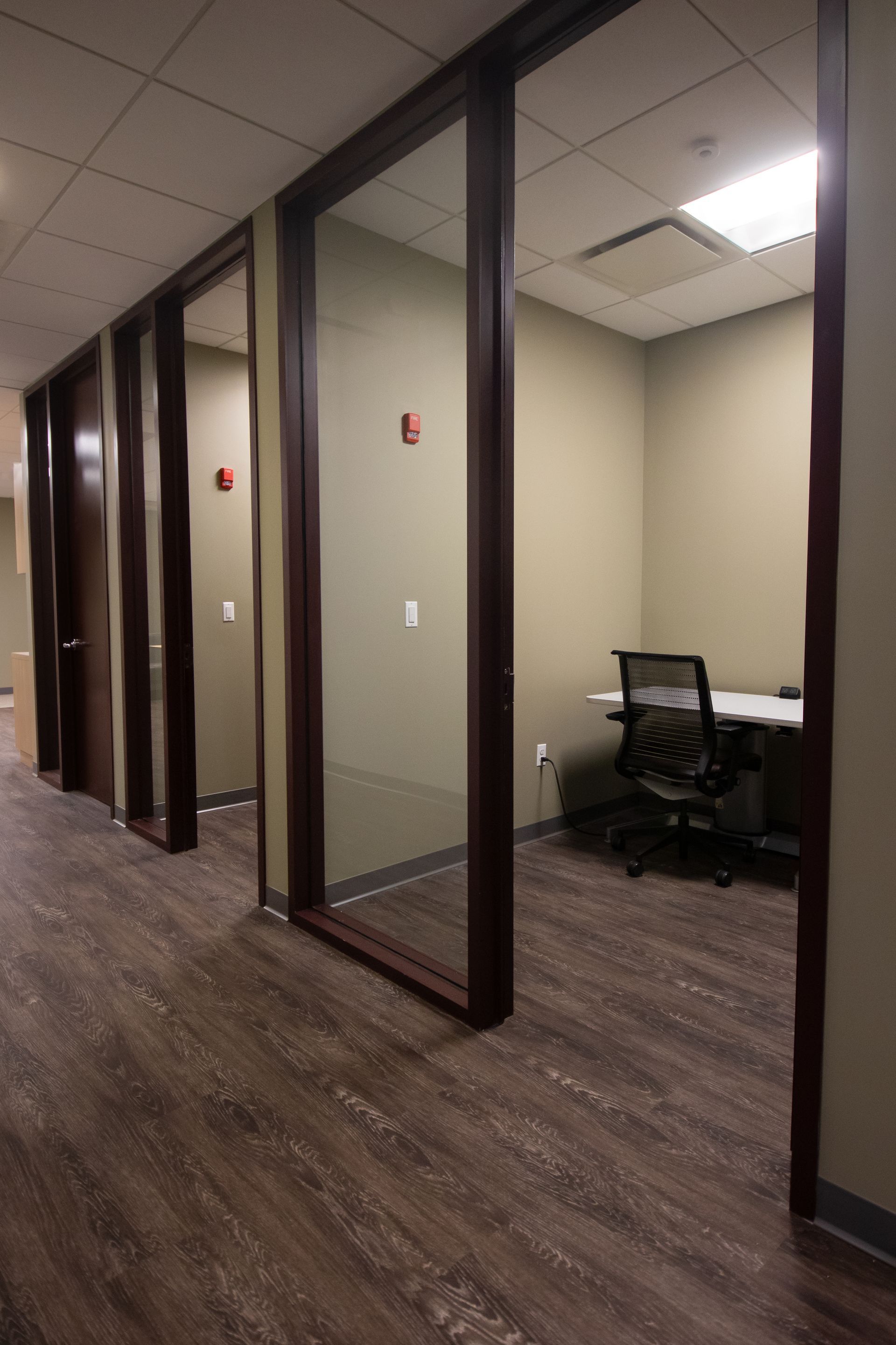 A hallway with small office rooms. One room is open, showing a desk, chair, and light.