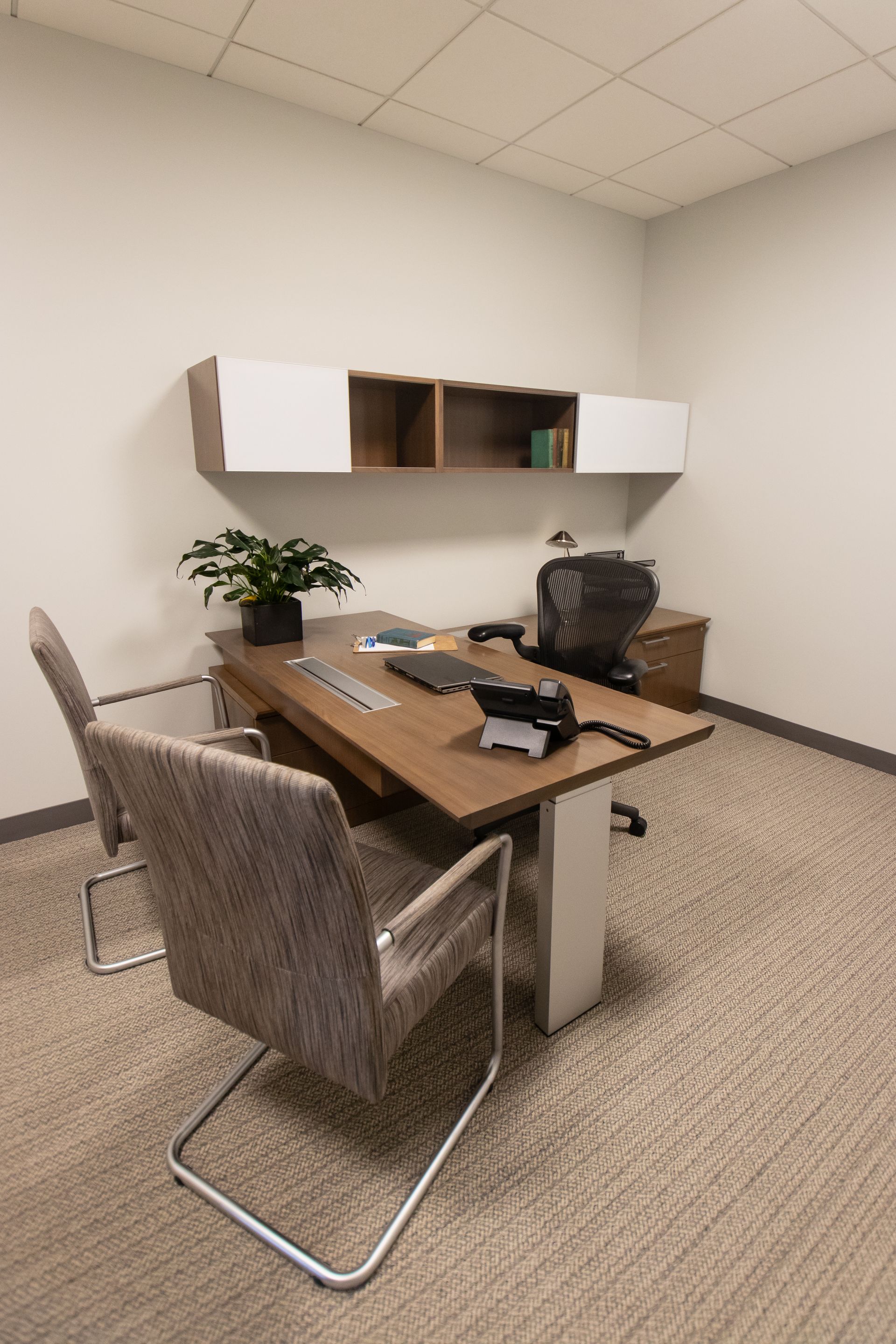 Office with desk, chairs, and overhead storage. Neutral colors. Carpeted floor.