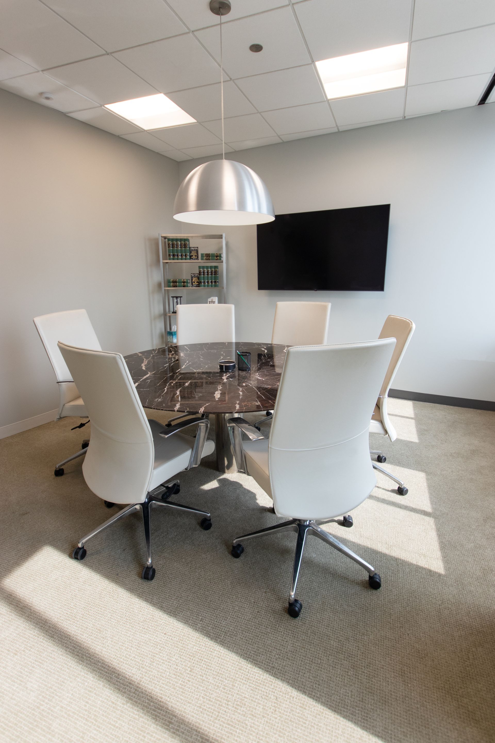 Conference room with white chairs around a brown table. A large screen and silver light fixture are present.