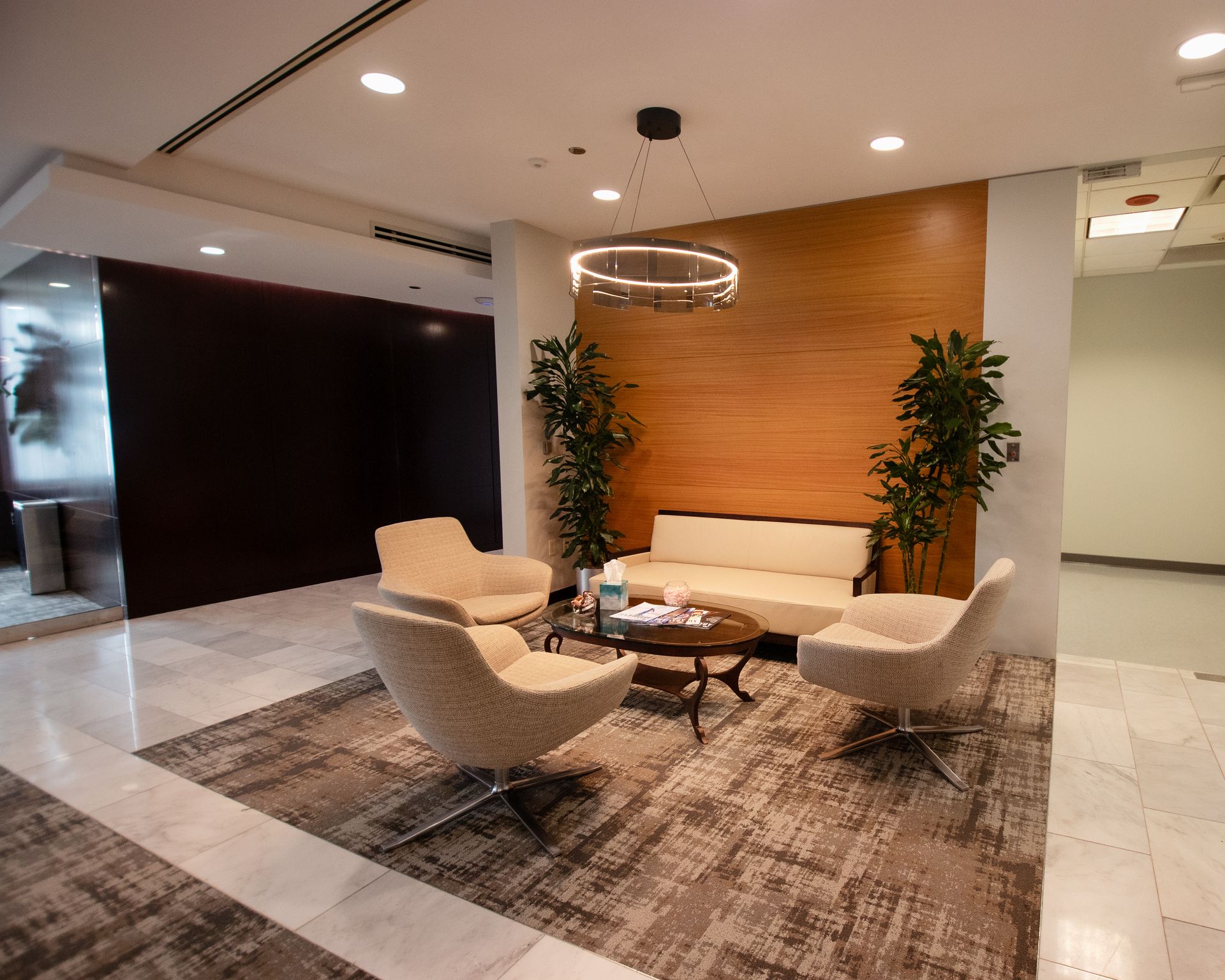 Lounge area with seating on a patterned rug; wood-paneled wall, plants, and a circular light fixture.
