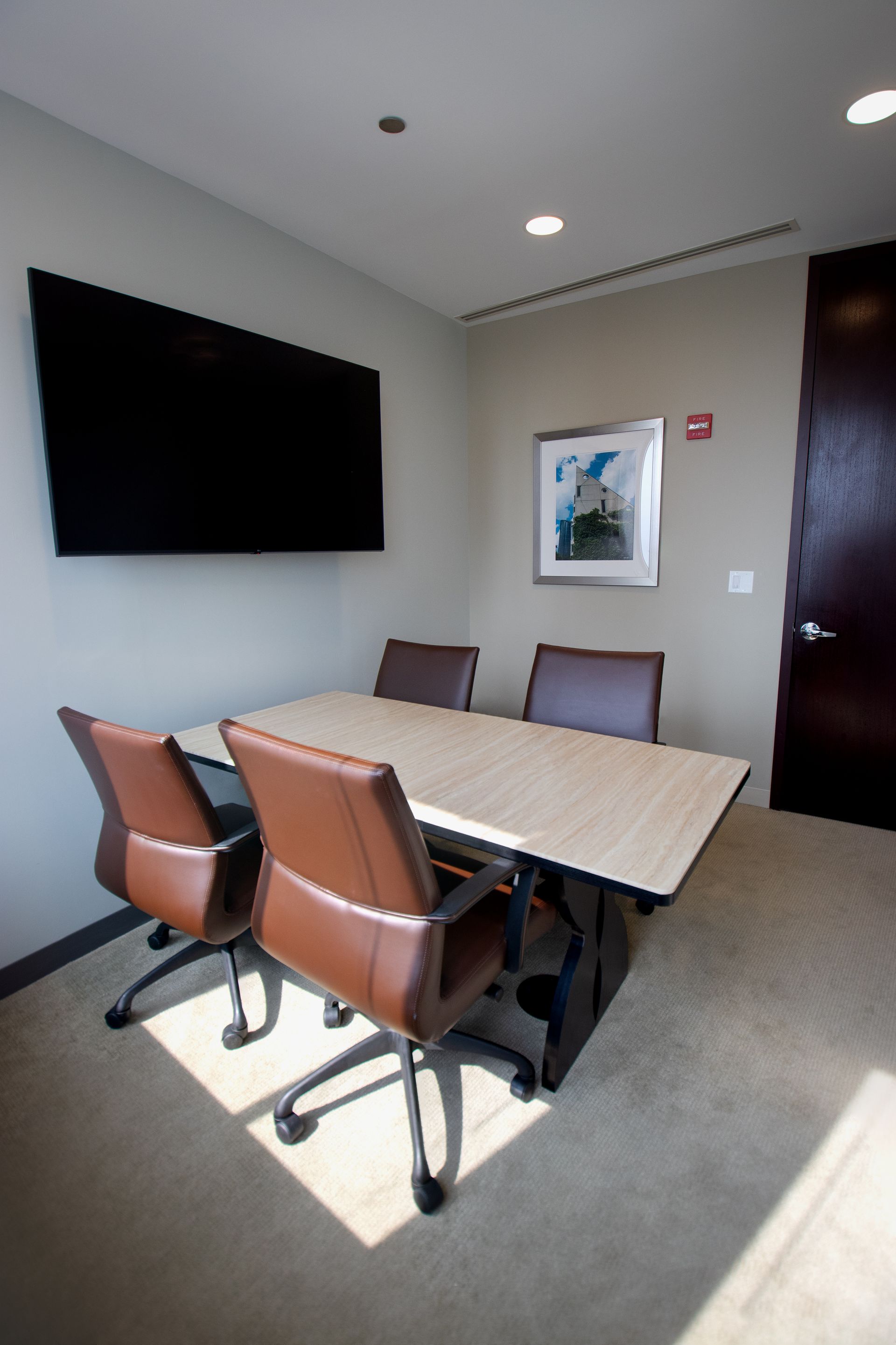 A small conference room with a rectangular table, four brown chairs, and a mounted TV.