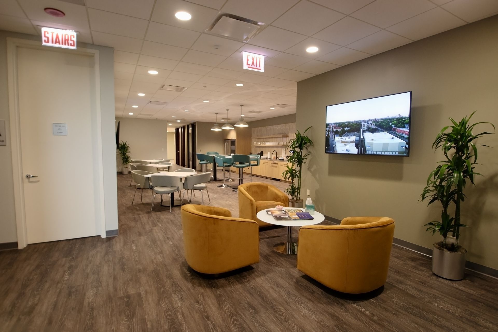 Modern office lounge area with yellow chairs, TV, tables, plants, and dining area.