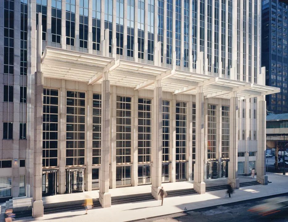 Office building facade with tall white columns, glass windows, and people walking on the sidewalk.