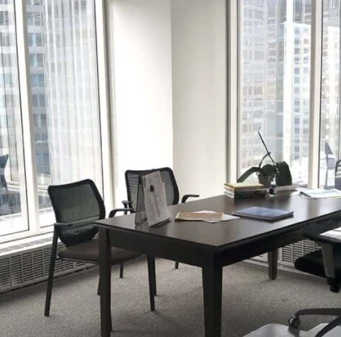 Office with large windows overlooking city buildings. Desk with chairs, papers, and plant.