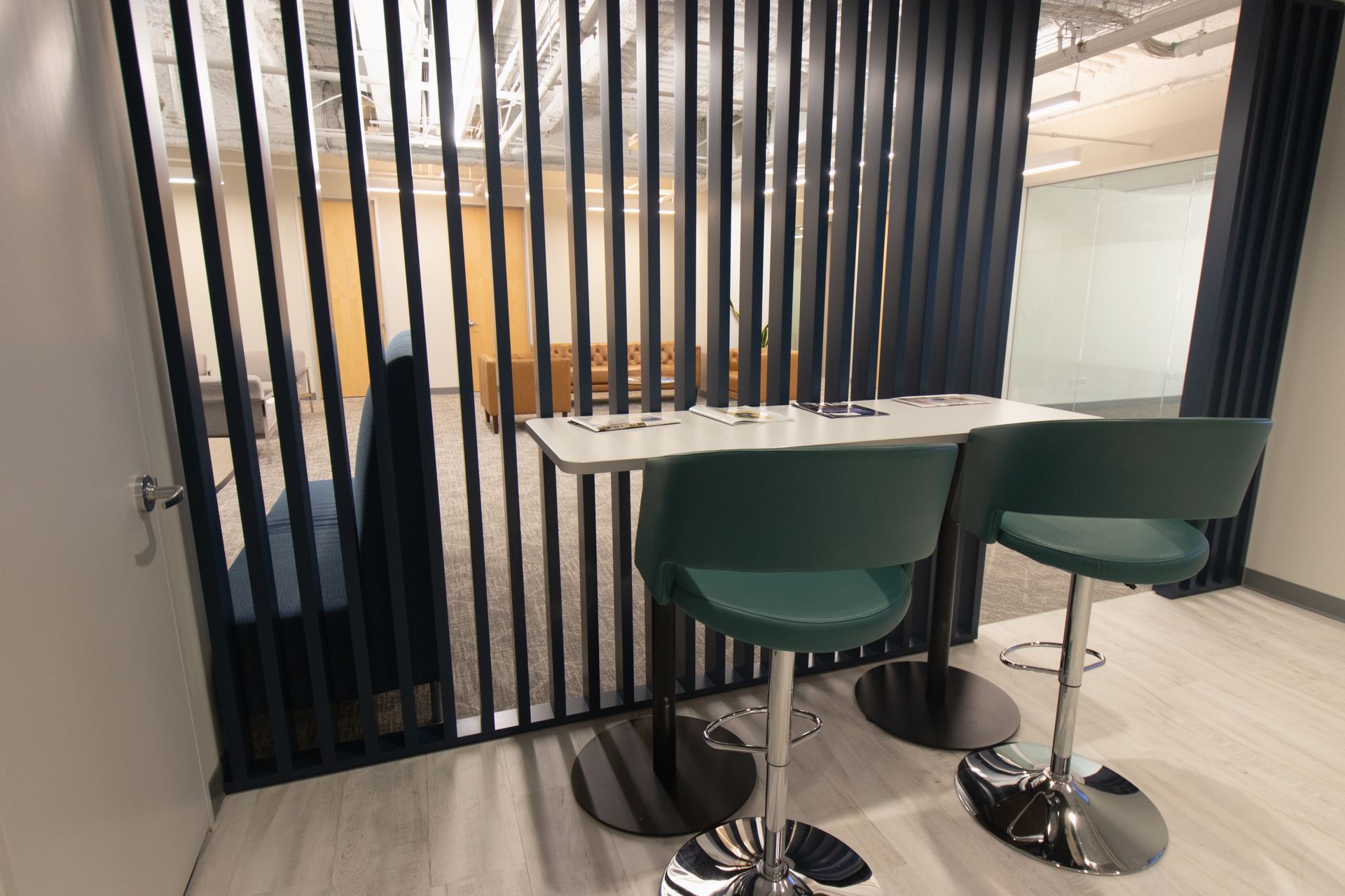 Two green bar stools sit in front of a white table, next to a black slat wall.