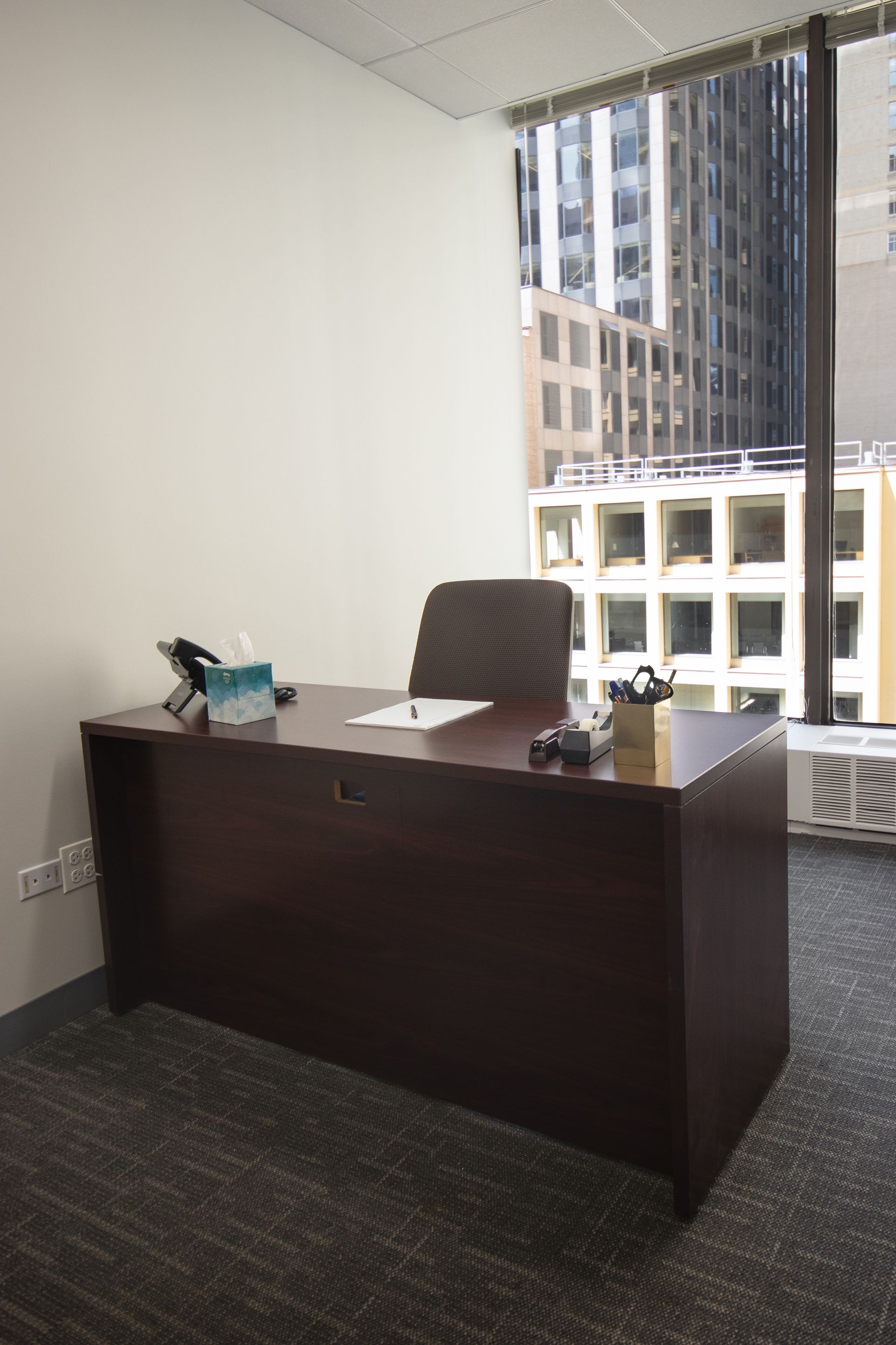 Office with desk, chair, window view of city buildings, dark carpet, white wall.