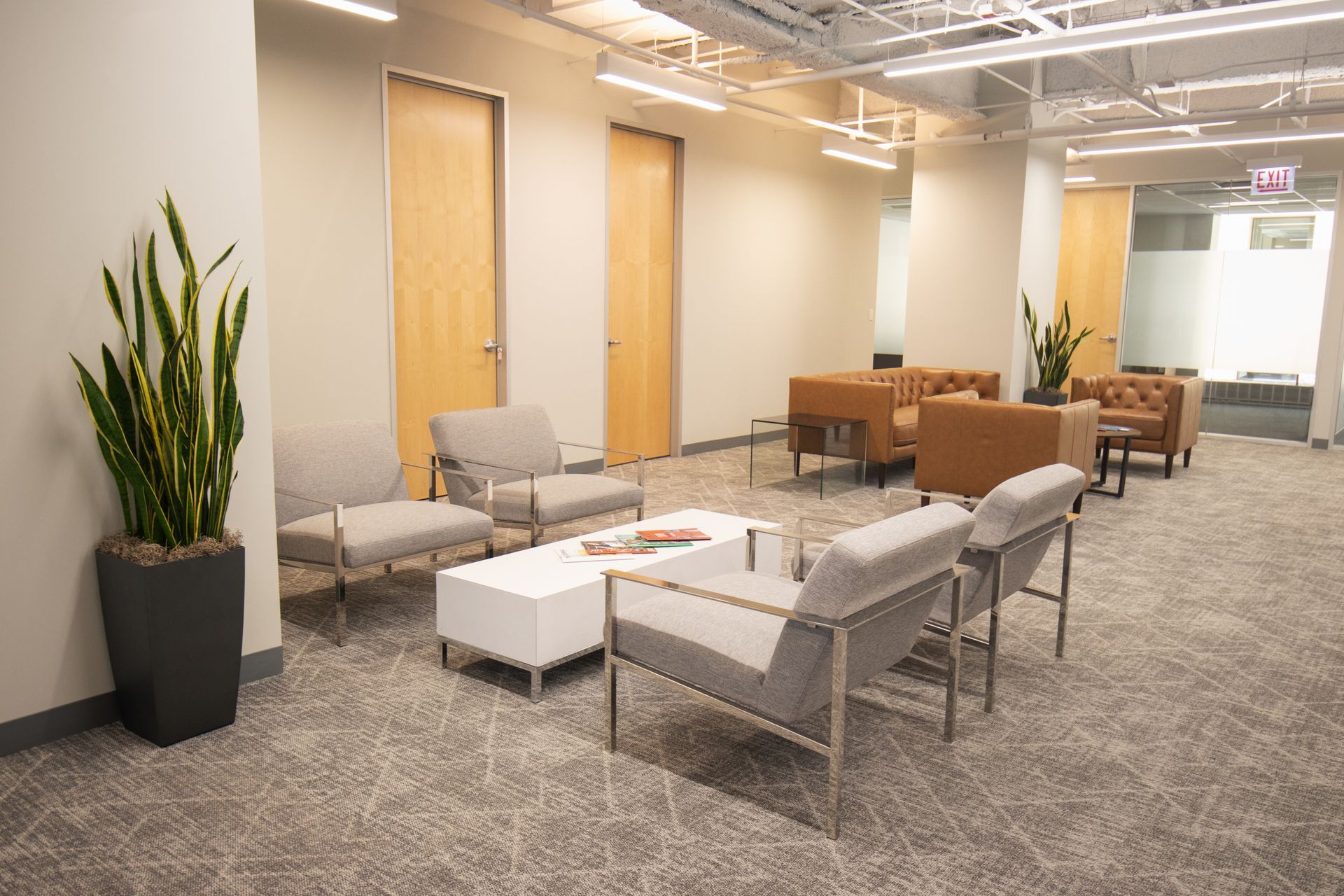 Lounge area with gray chairs, a white coffee table, and tall potted plant. Two brown leather sofas in the background.
