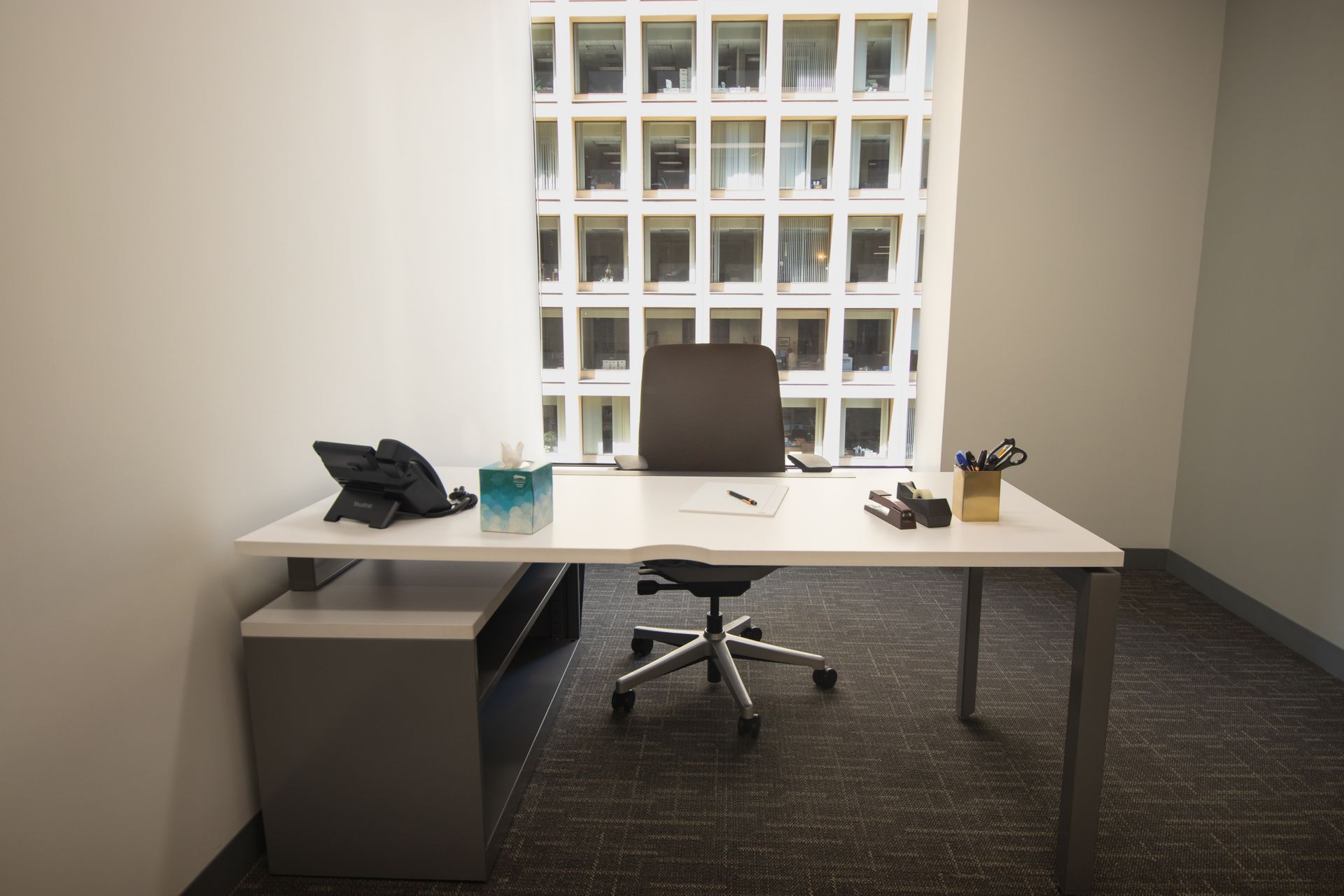 Office desk with chair, phone, and supplies, in front of a window overlooking a building.