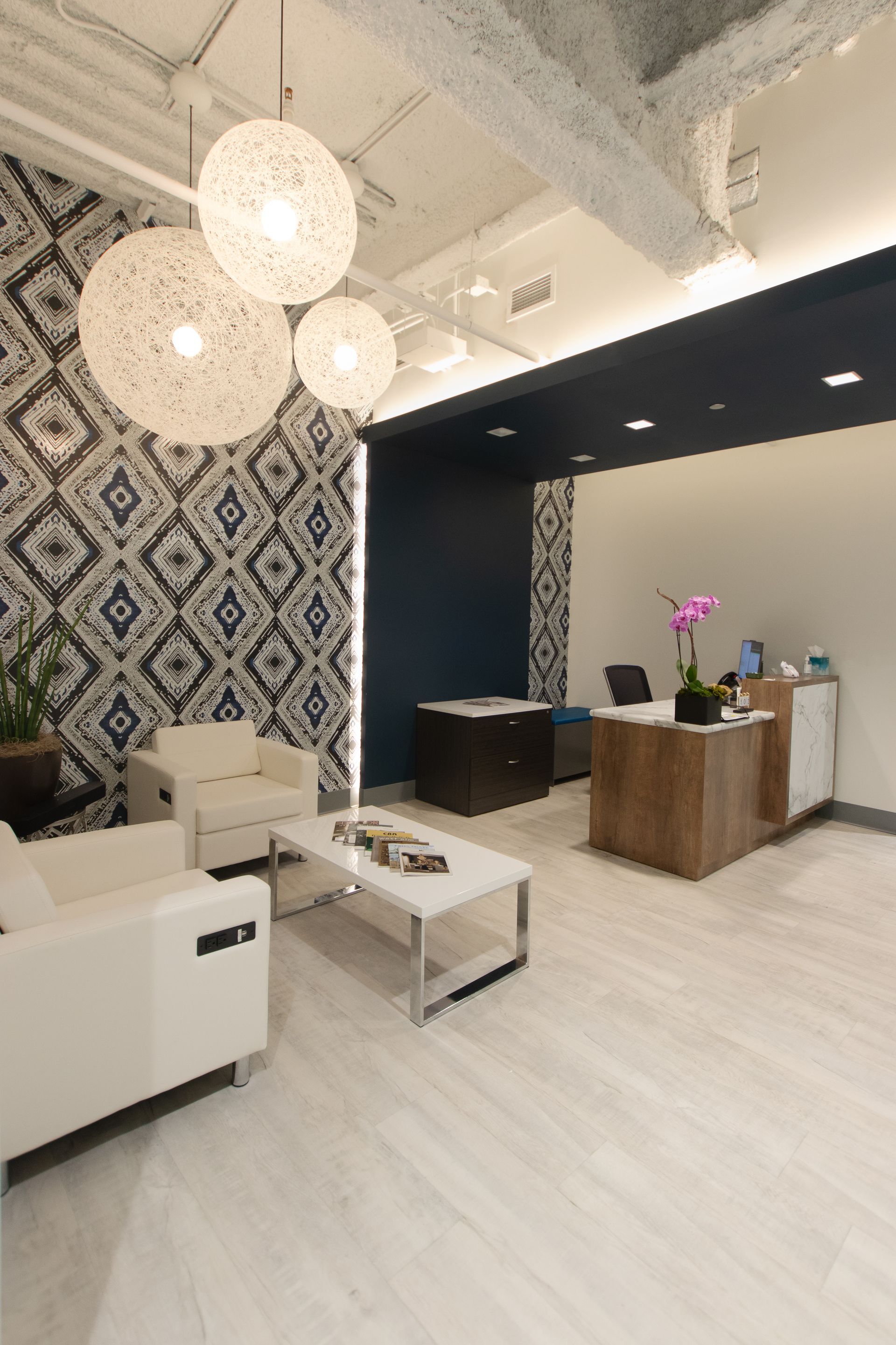 Modern office reception area with patterned wall, white seating, globe lights, and wooden reception desk.
