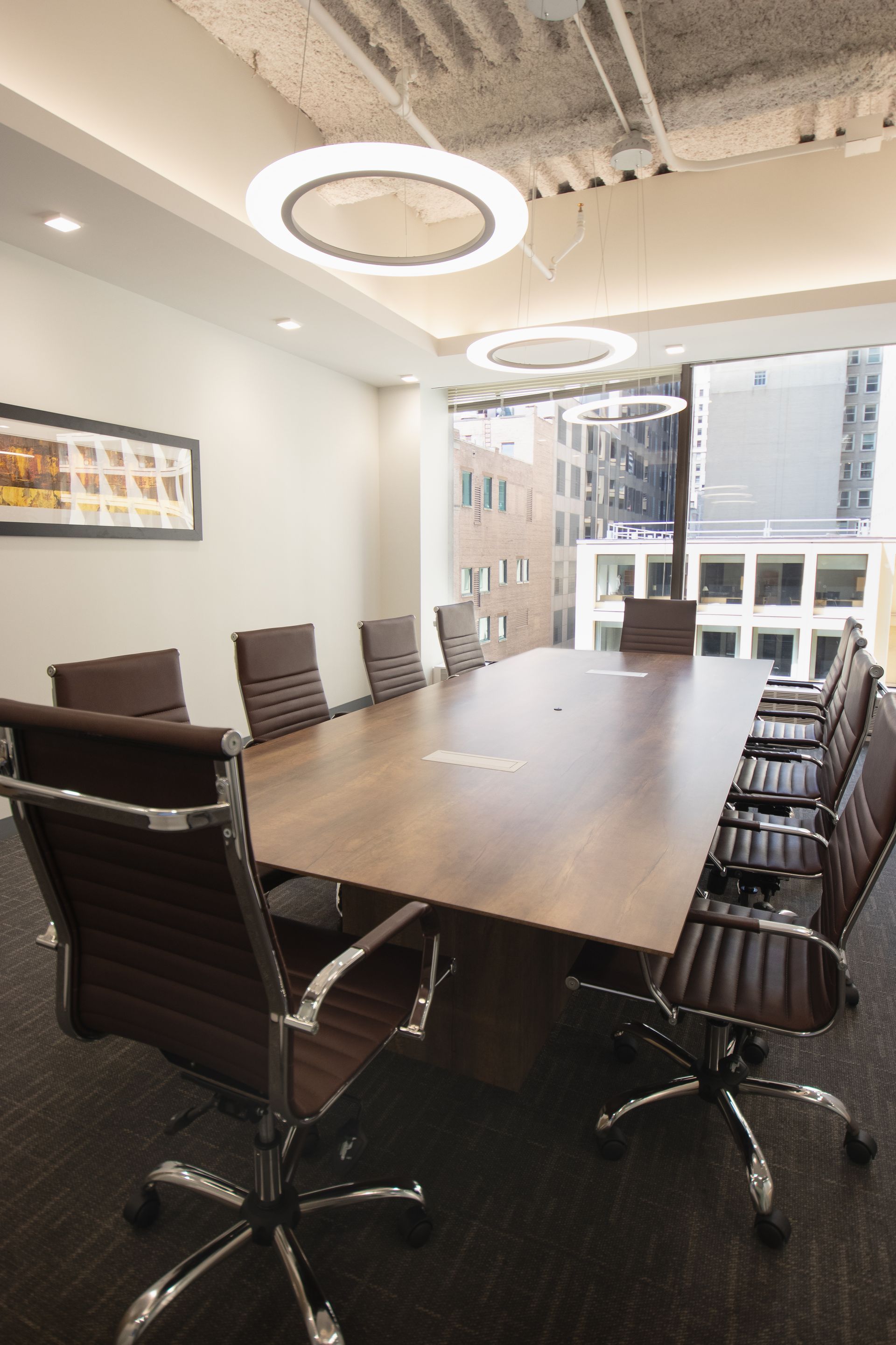 Conference room with long wooden table, brown leather chairs, and city view through large window.