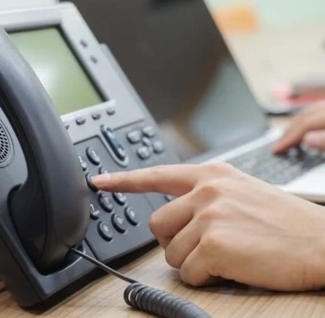 Person pressing a button on a black office phone, laptop in the background.