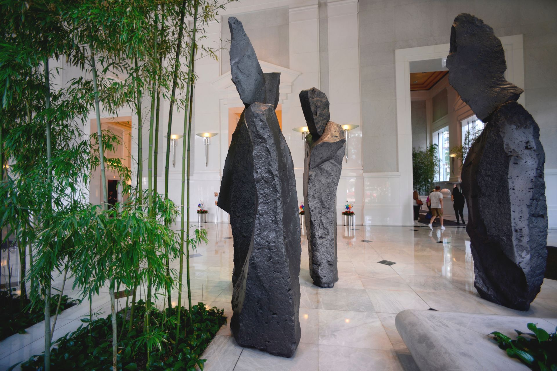 Large dark stone sculptures in a bright white room with bamboo plants. People are in the background.