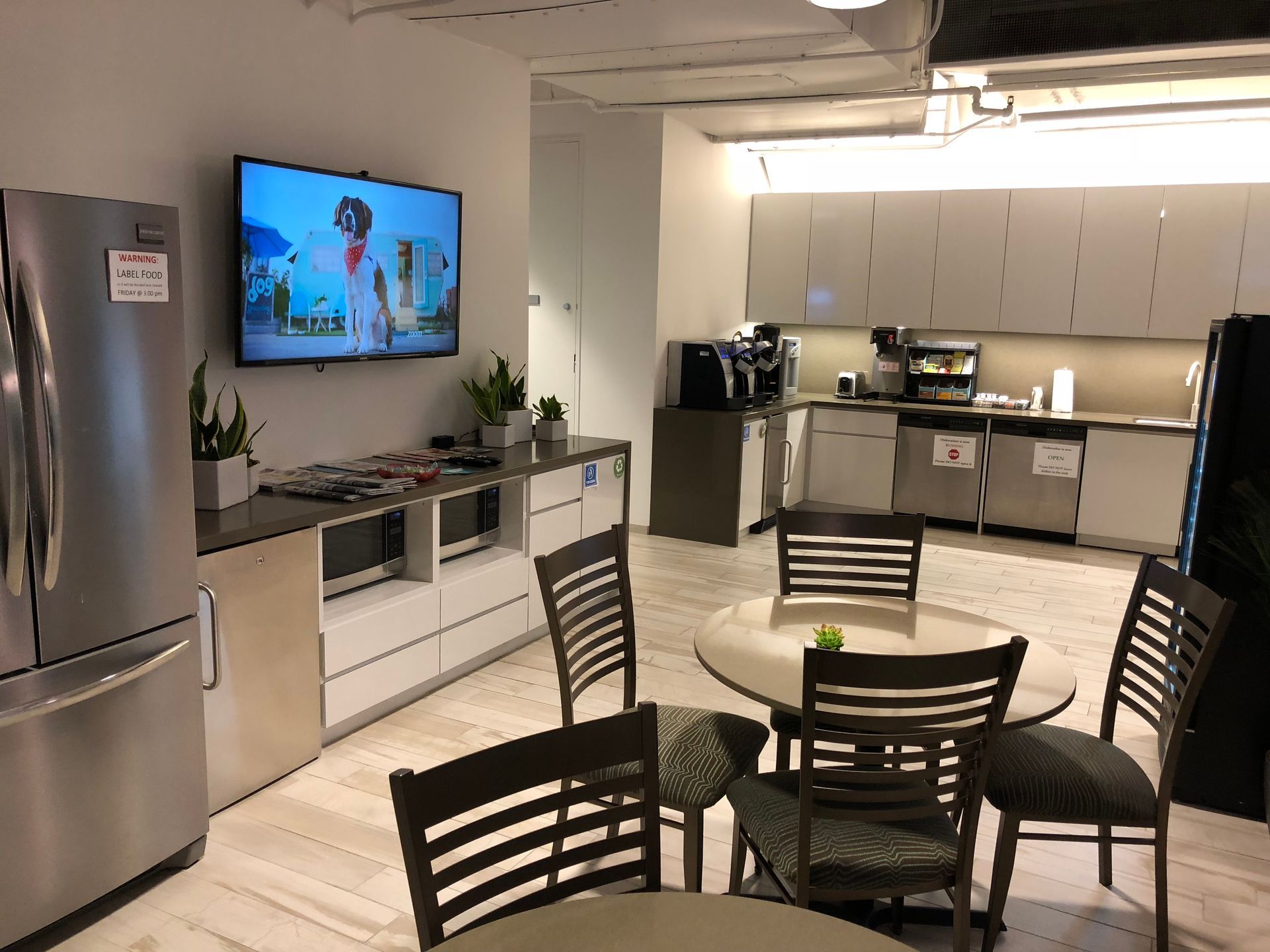 Office break room with stainless steel refrigerator, television, microwave, and coffee maker. Round tables with chairs.