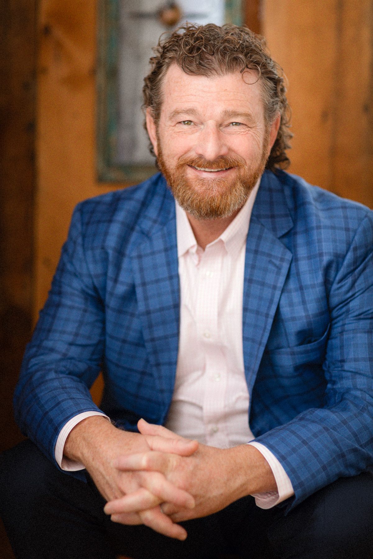 Man with curly brown hair and beard, wearing a blue plaid blazer, smiling.