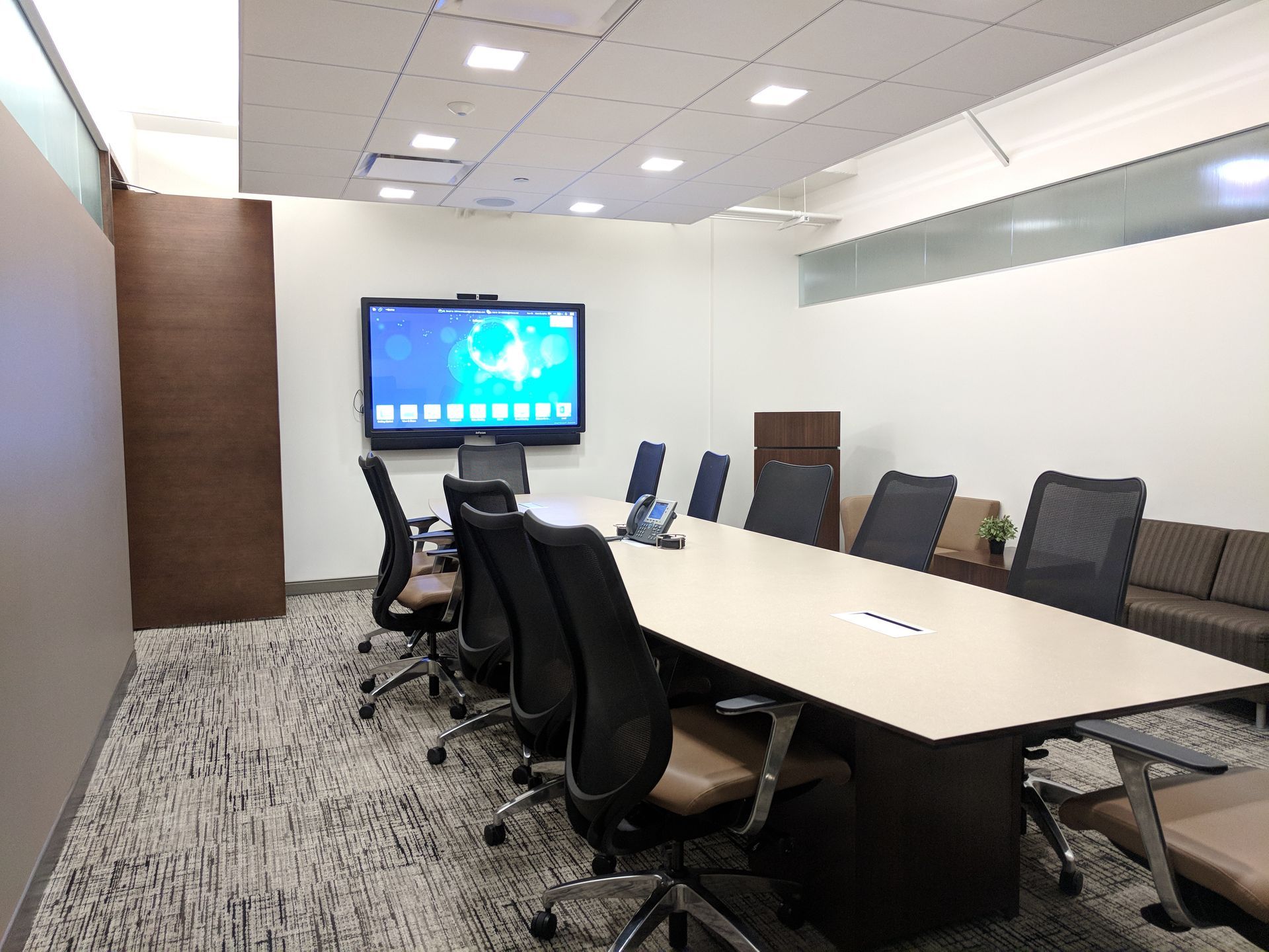 Conference room with large screen, table, chairs, neutral colors, and patterned carpet.