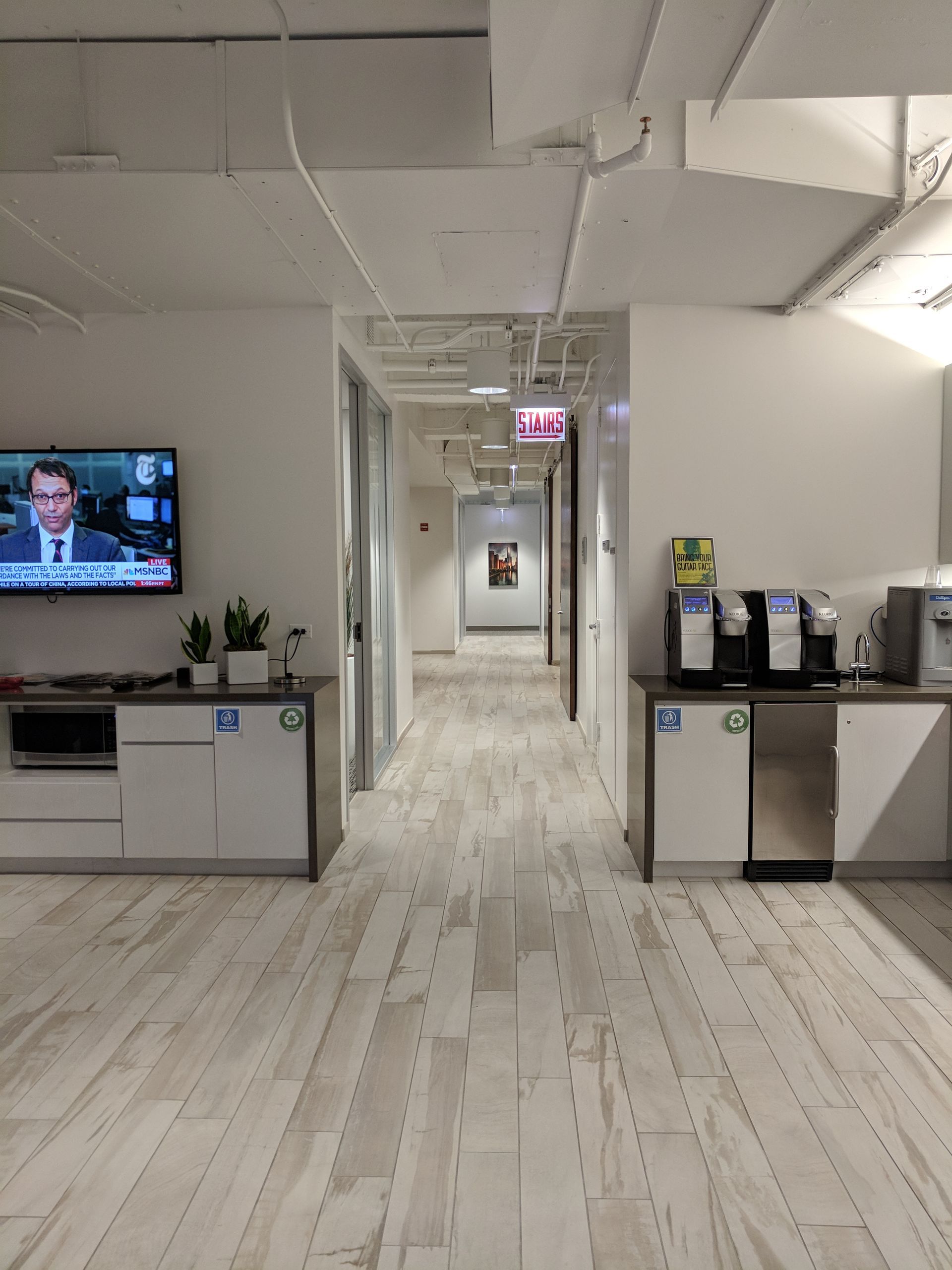 Office hallway with coffee stations on either side, television at left, white walls and wood floor.