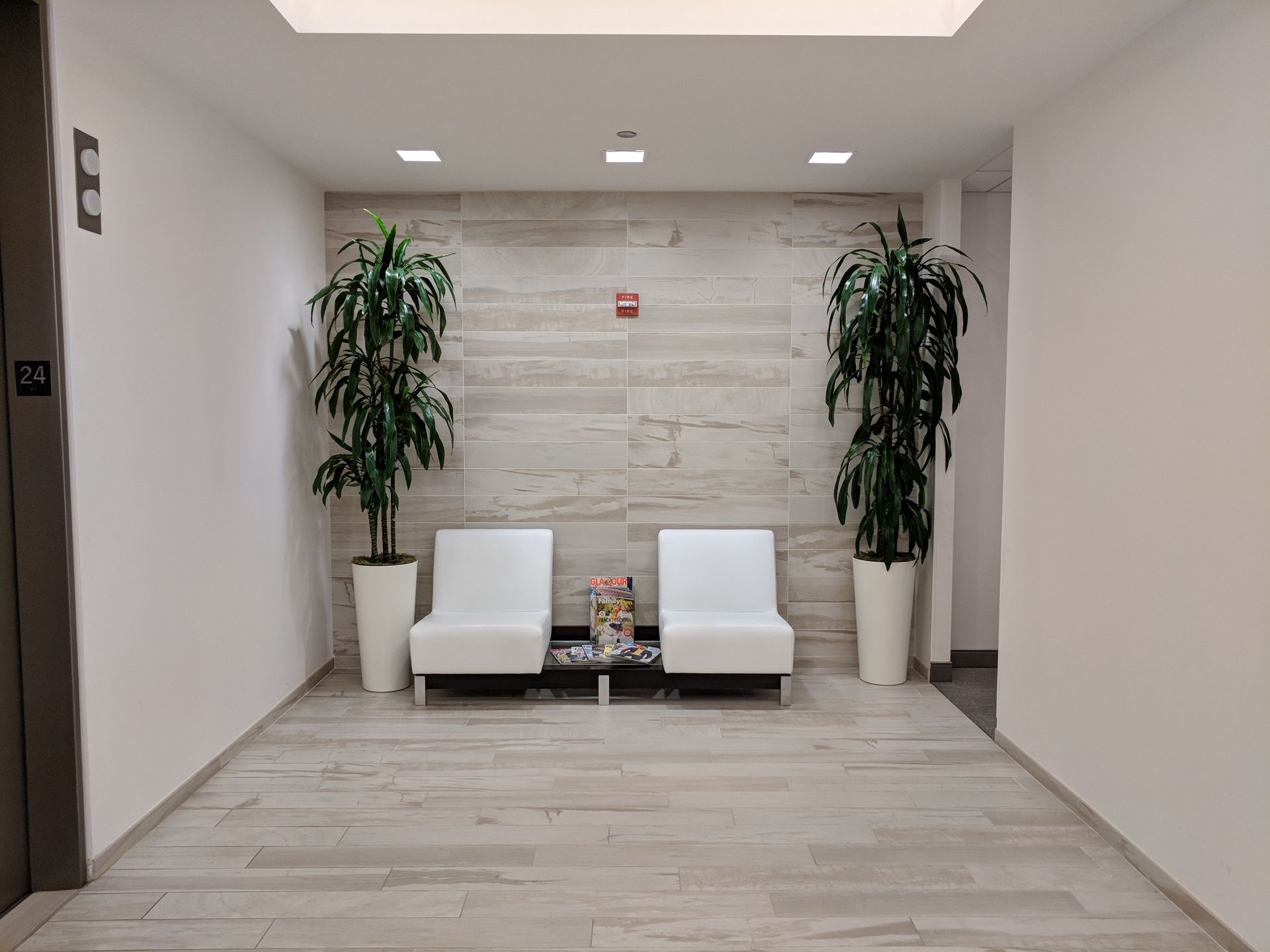 Hallway with two white chairs, two tall plants, wood-like tile, and an elevator.