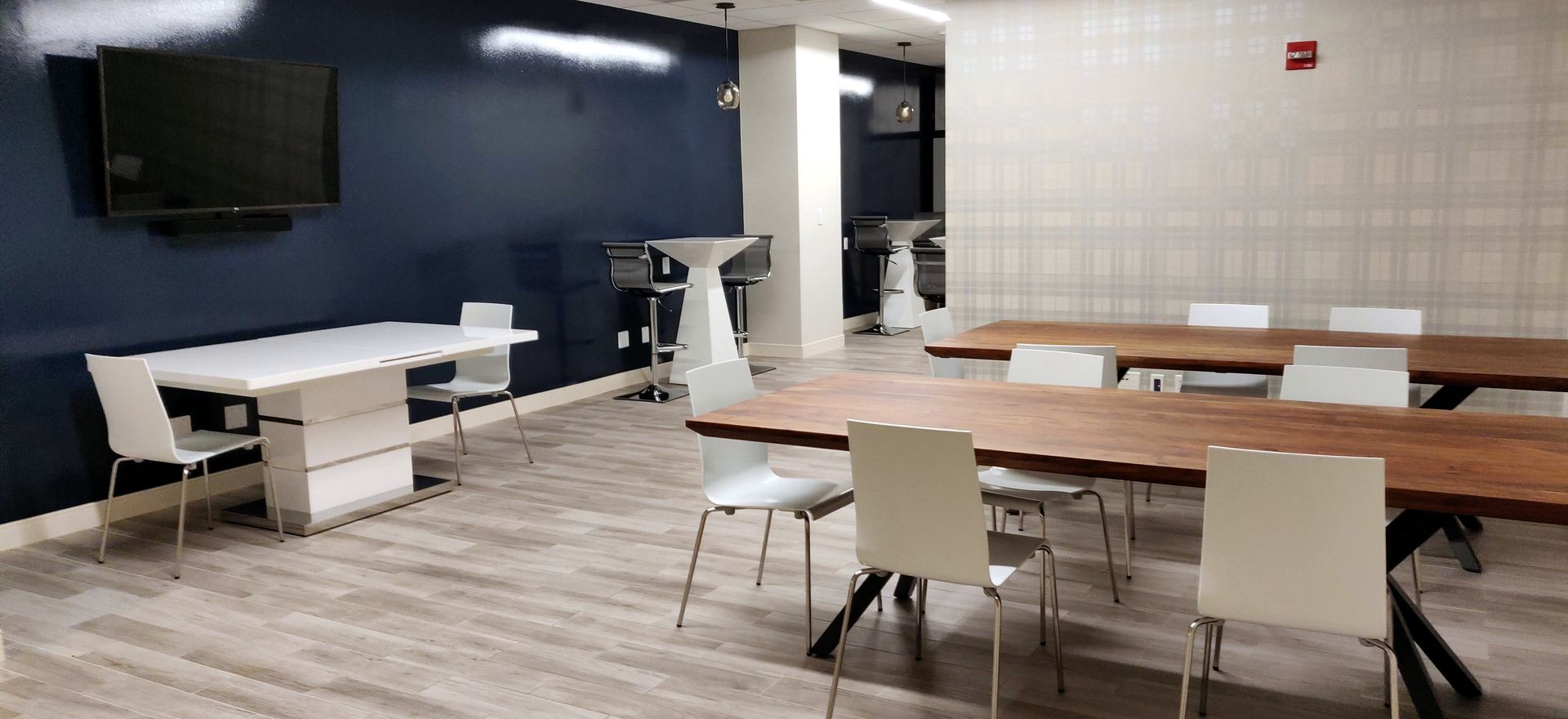 An empty, modern meeting room with tables and white chairs. Dark blue wall with a mounted TV.