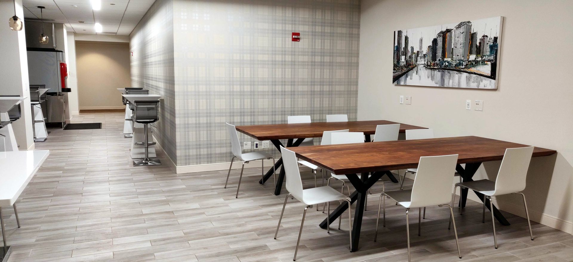 Office break room with tables, chairs, and artwork; a hallway extends to the left.
