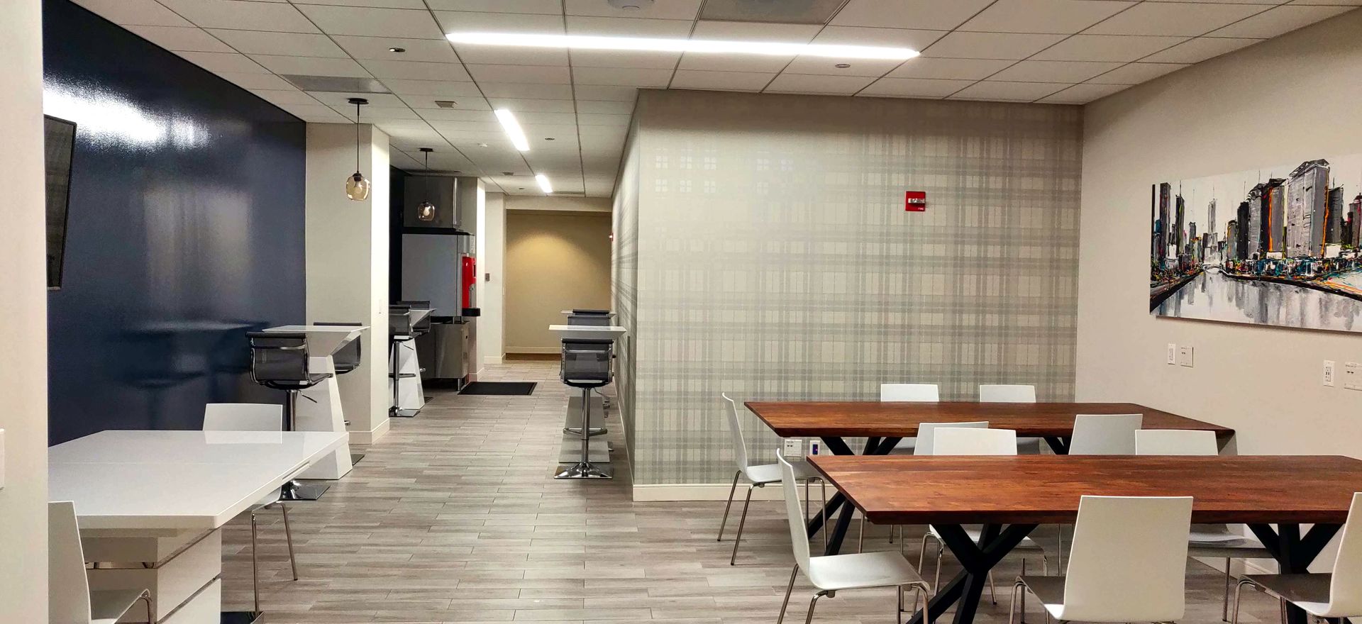Office breakroom with tables, chairs, and kitchenette. Dark blue wall, neutral wall, city art.