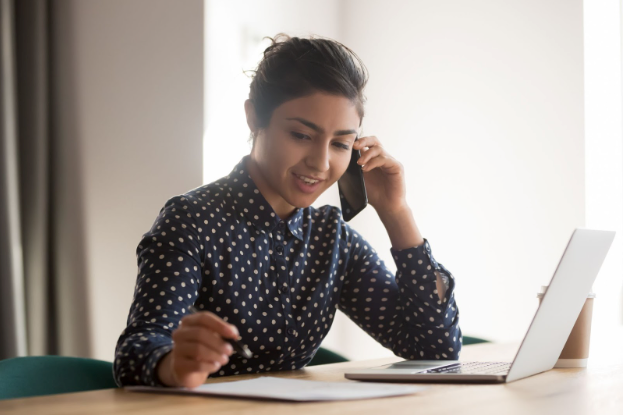 Woman on phone, smiles, looking at laptop and paperwork at a desk.