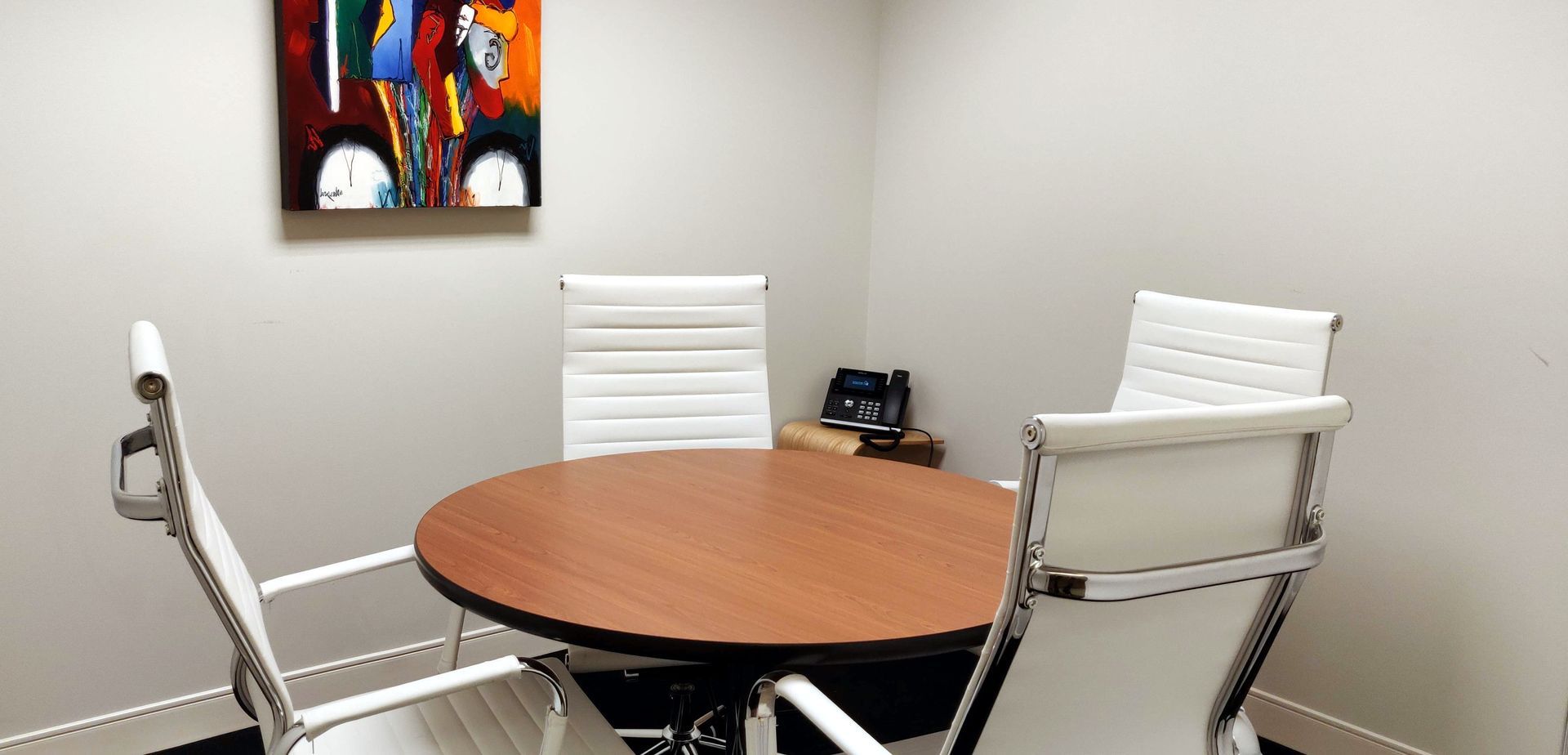 A small conference room with a round table and three white chairs, and abstract art on the wall.