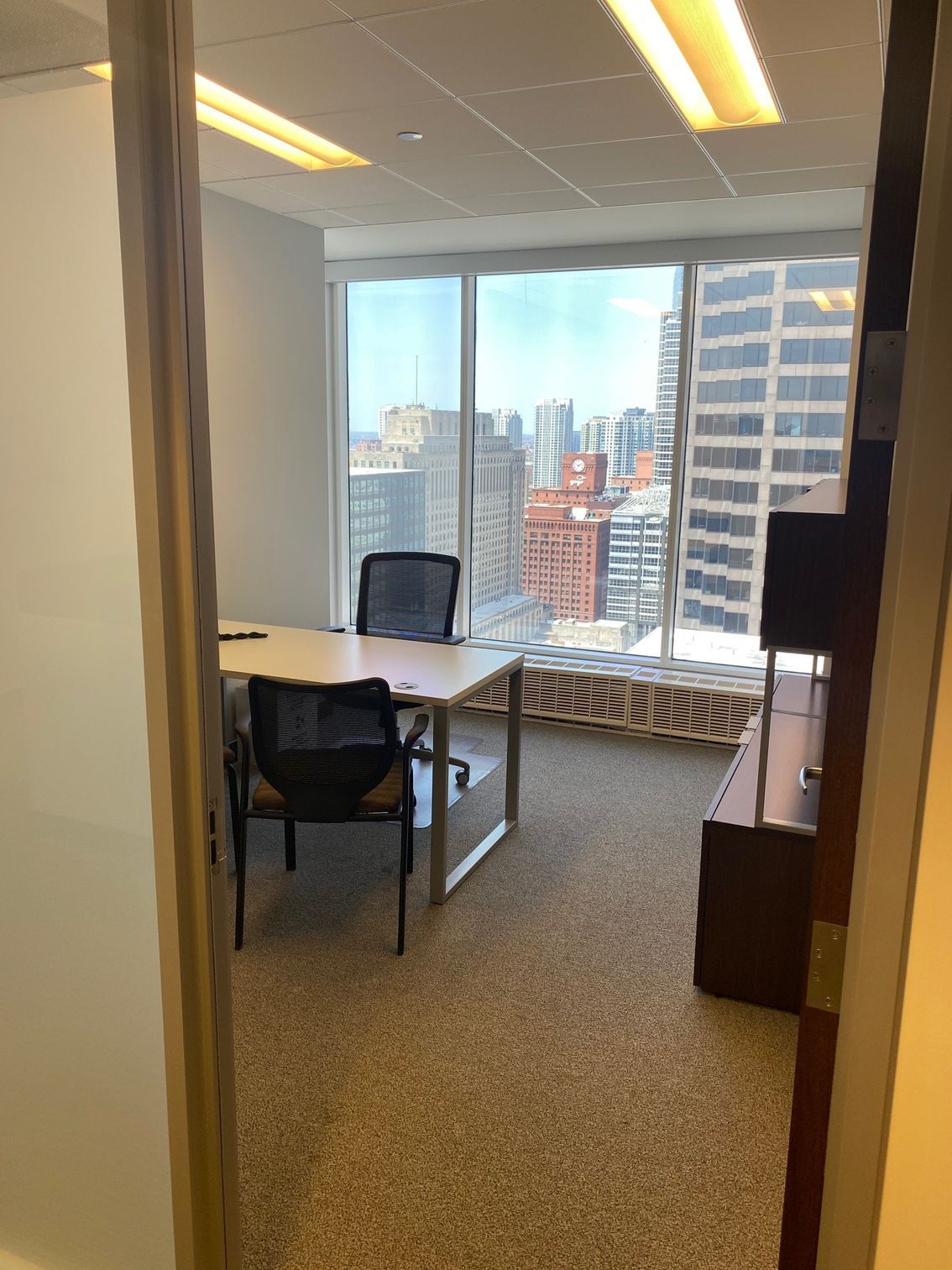 Office interior with desk, two chairs, and city view through a large window.