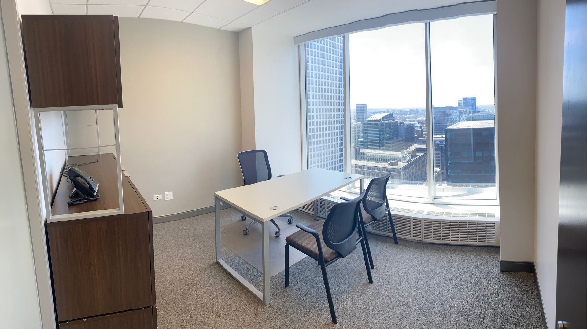 Office with desk and chairs facing a large window overlooking a cityscape. Brown cabinet on left.