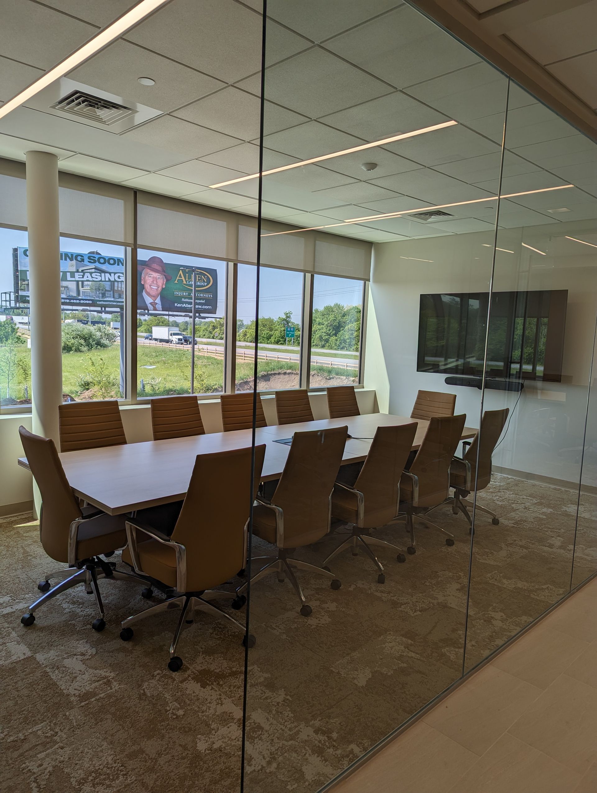 Conference room with a large table, chairs, and windows overlooking greenery. A TV is mounted on the wall.