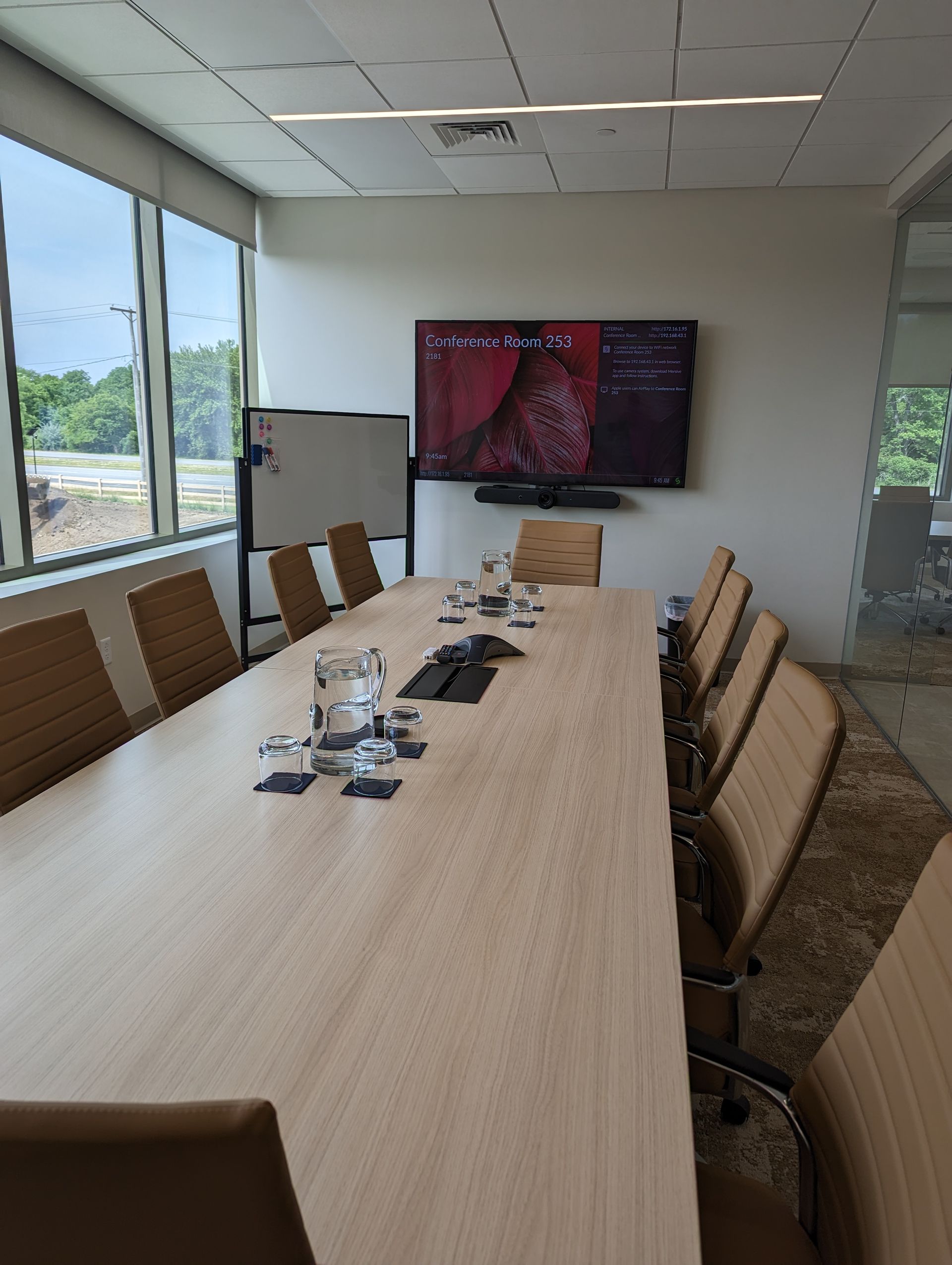 Conference room with long table, chairs, and mounted TV. Windows on the left, whiteboard, and water glasses on the table.