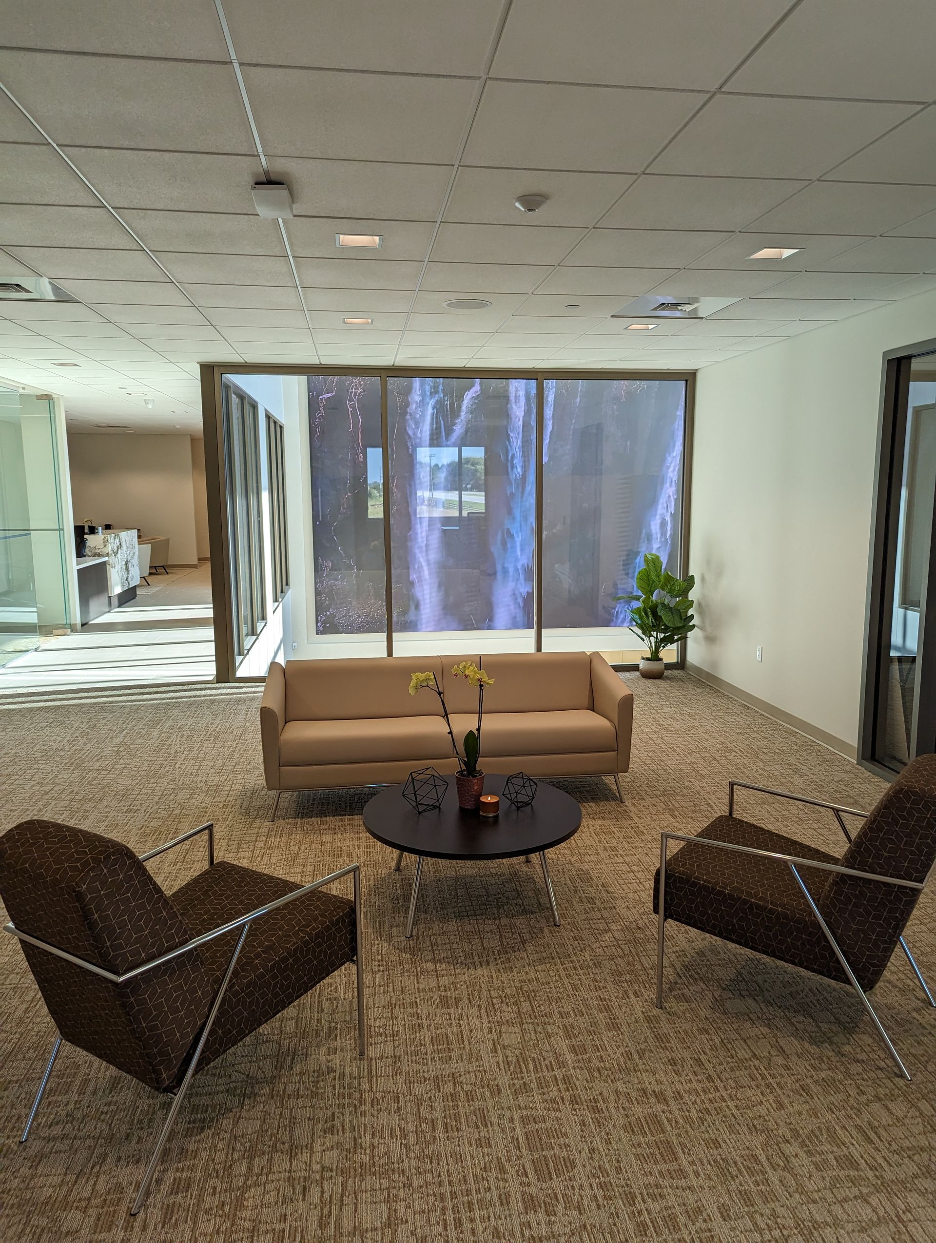 Lounge area with sofa, chairs, round table, and water feature backdrop. Carpeted floor, white walls, and ceiling.
