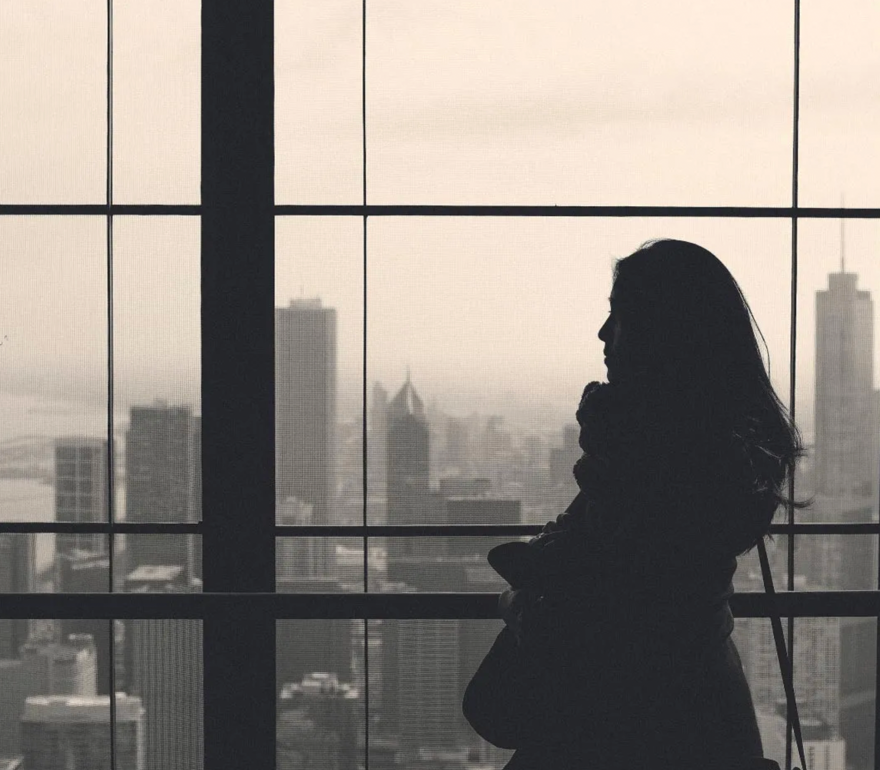 Silhouette of person looking out a large window at a cityscape with tall buildings.