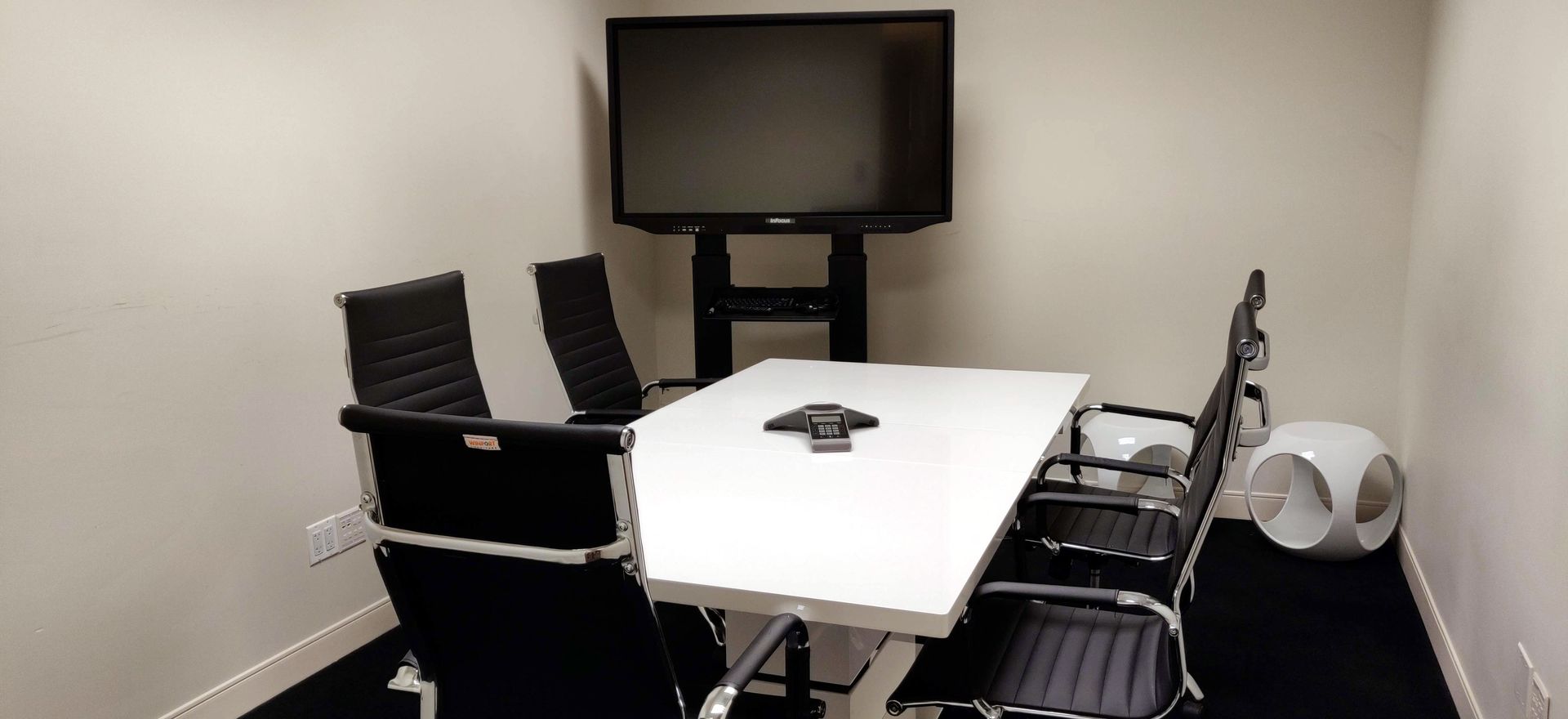 A small meeting room with a white table, black chairs, and a large TV.